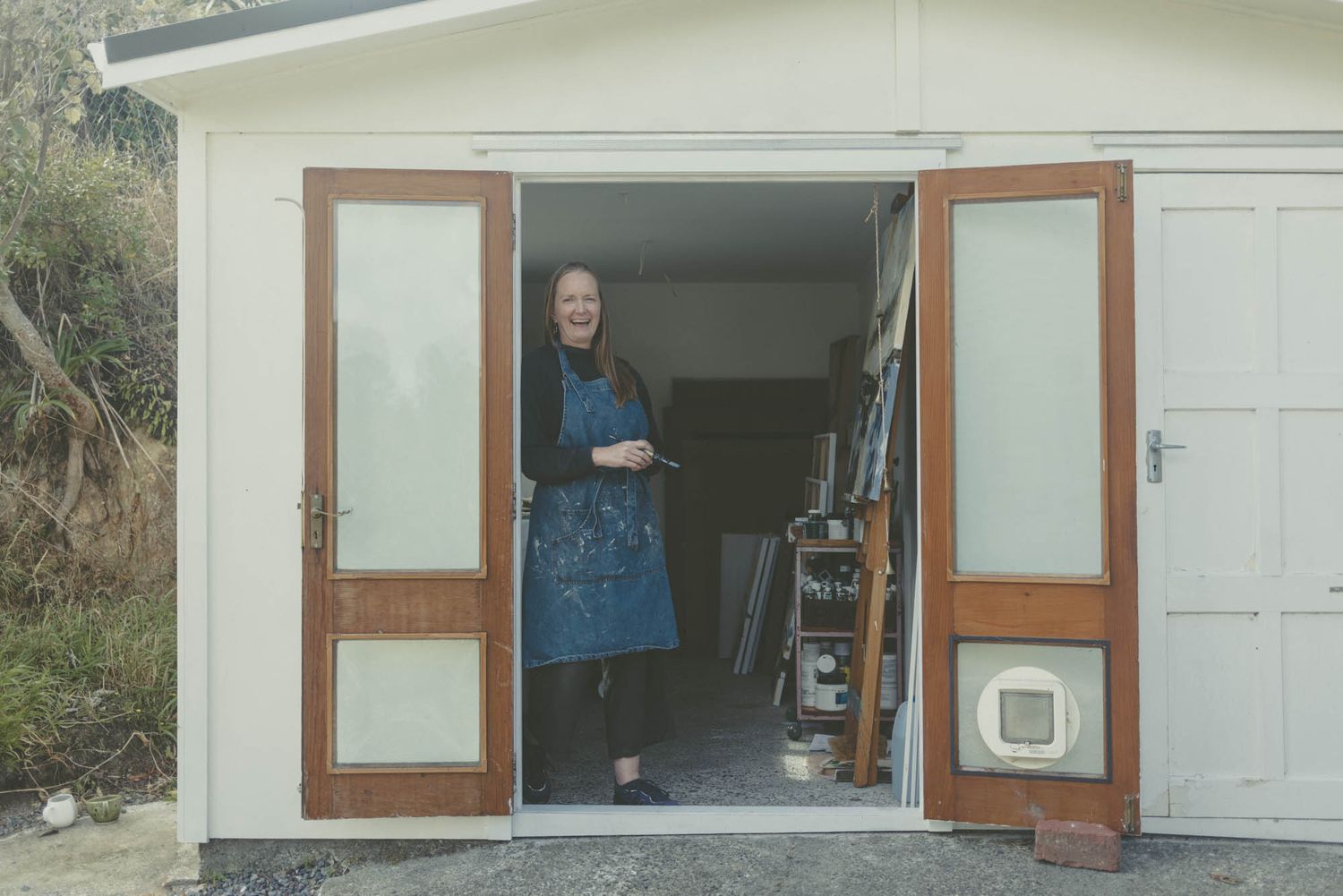Artist standing in doorway of white studio building with wooden doors.