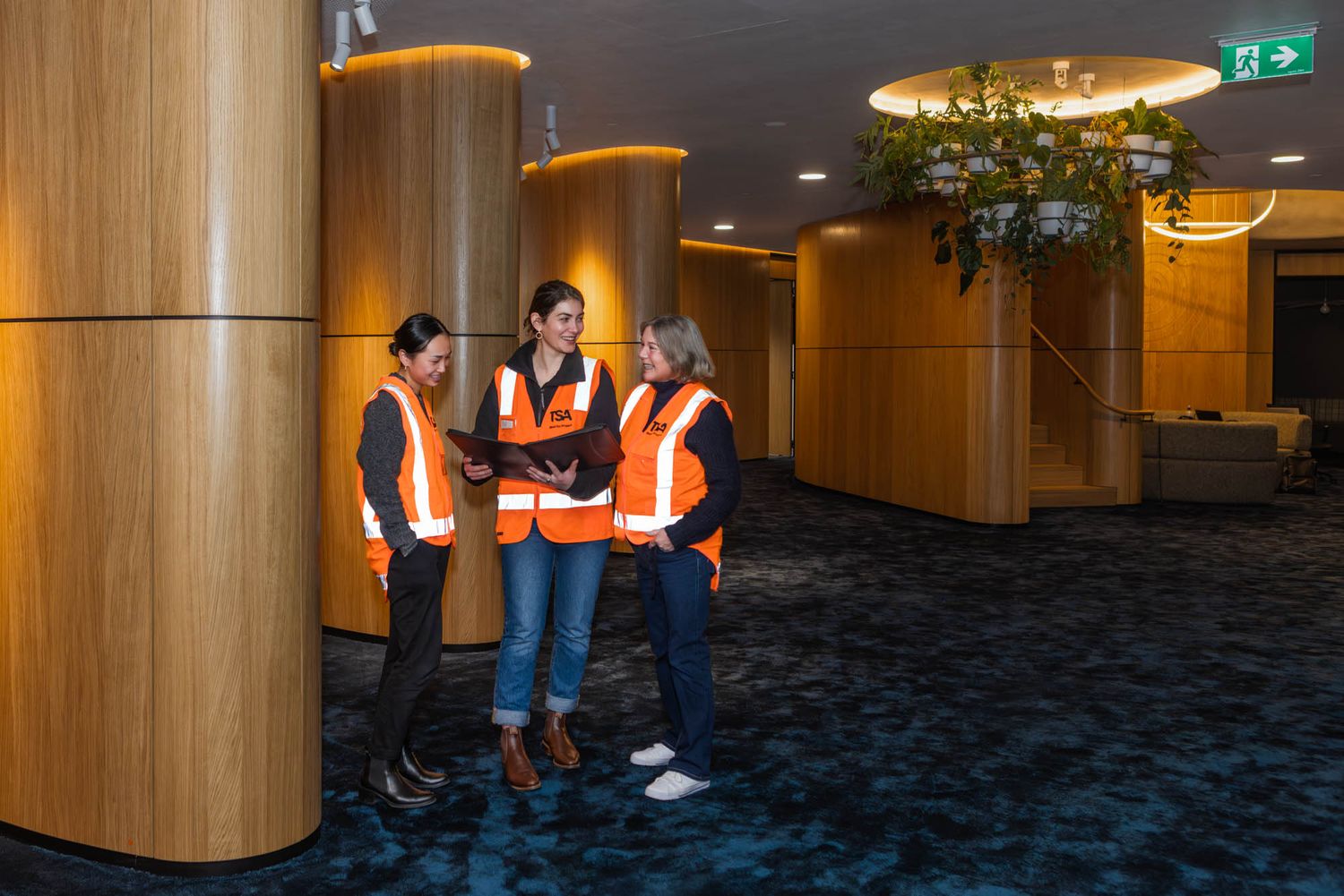 Modern office hallway with blue carpeting, wooden columns, and safety-vested workers visible in the corridor.