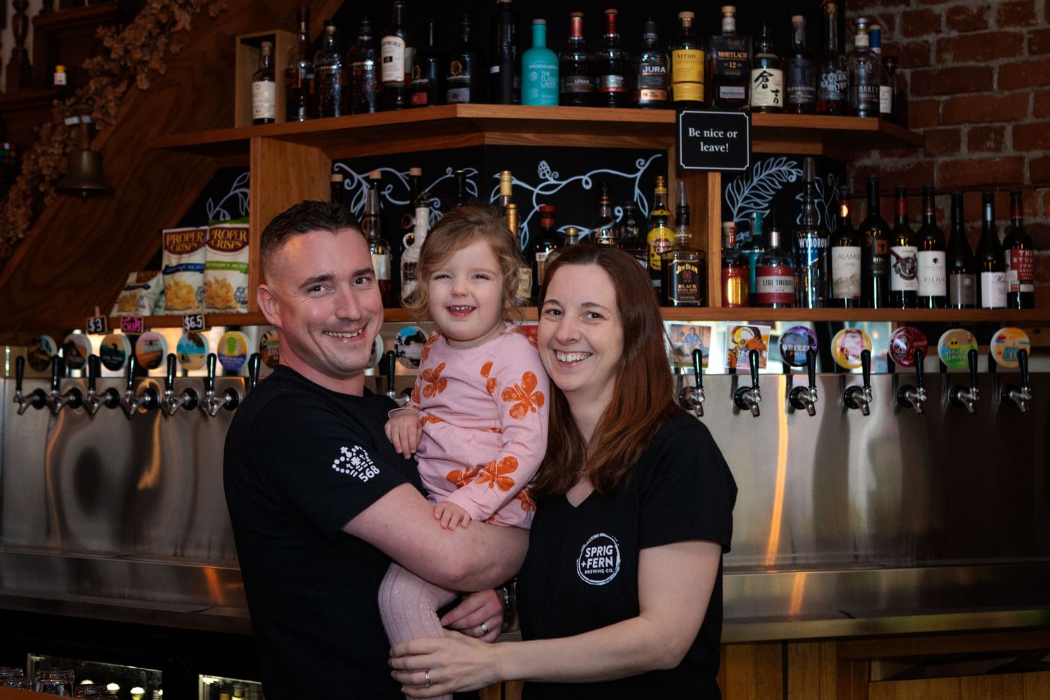 Family standing together behind Sprig and Fern bar counter with beer taps and bottles displayed.