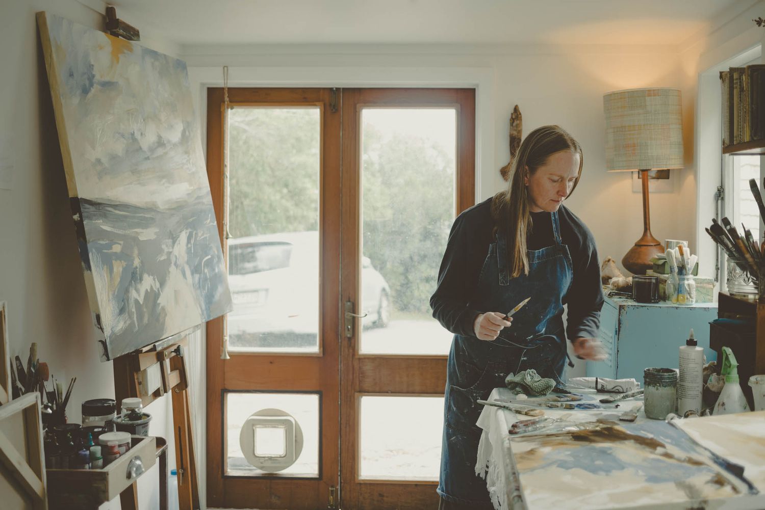 Artist working on a painting in a studio with glass doors and natural lighting.