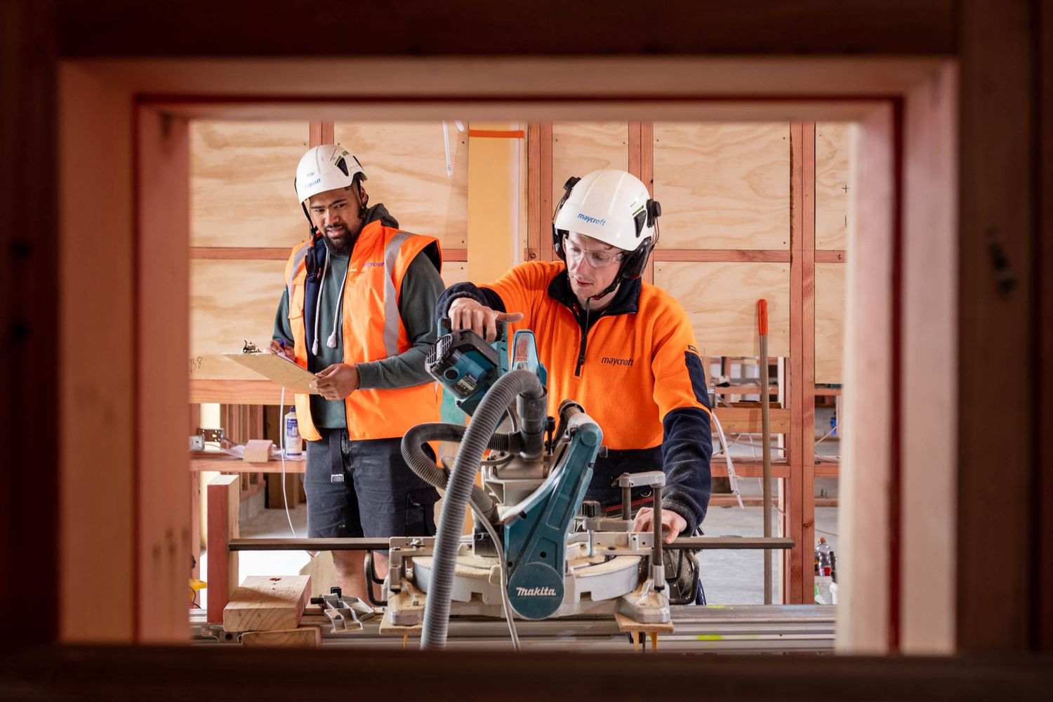 Two construction workers in orange hi-vis gear and white helmets using a circular saw inside a timber-framed building.