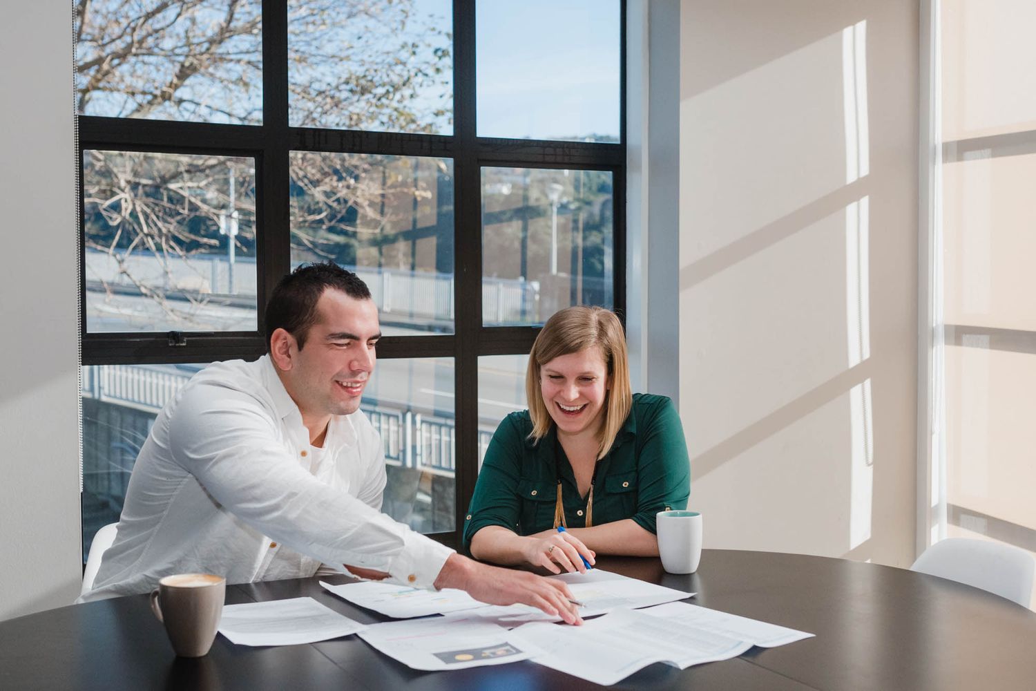 Two colleagues review paperwork together at a bright modern office desk near large windows.