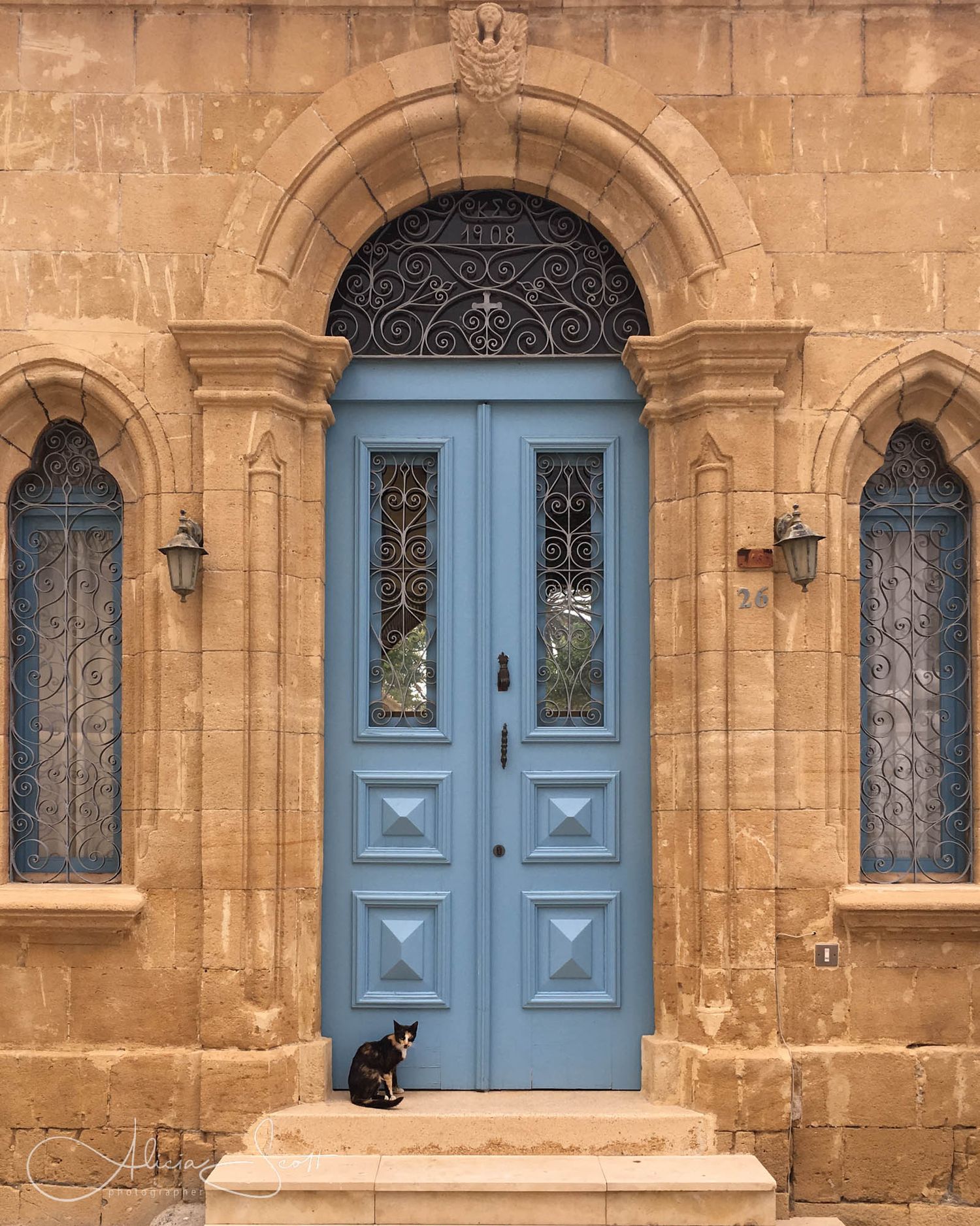 A tortieshell cat patiently sits before a blue double door with ornate ironwork on a tan stone building in Larnaca, Cyprus.