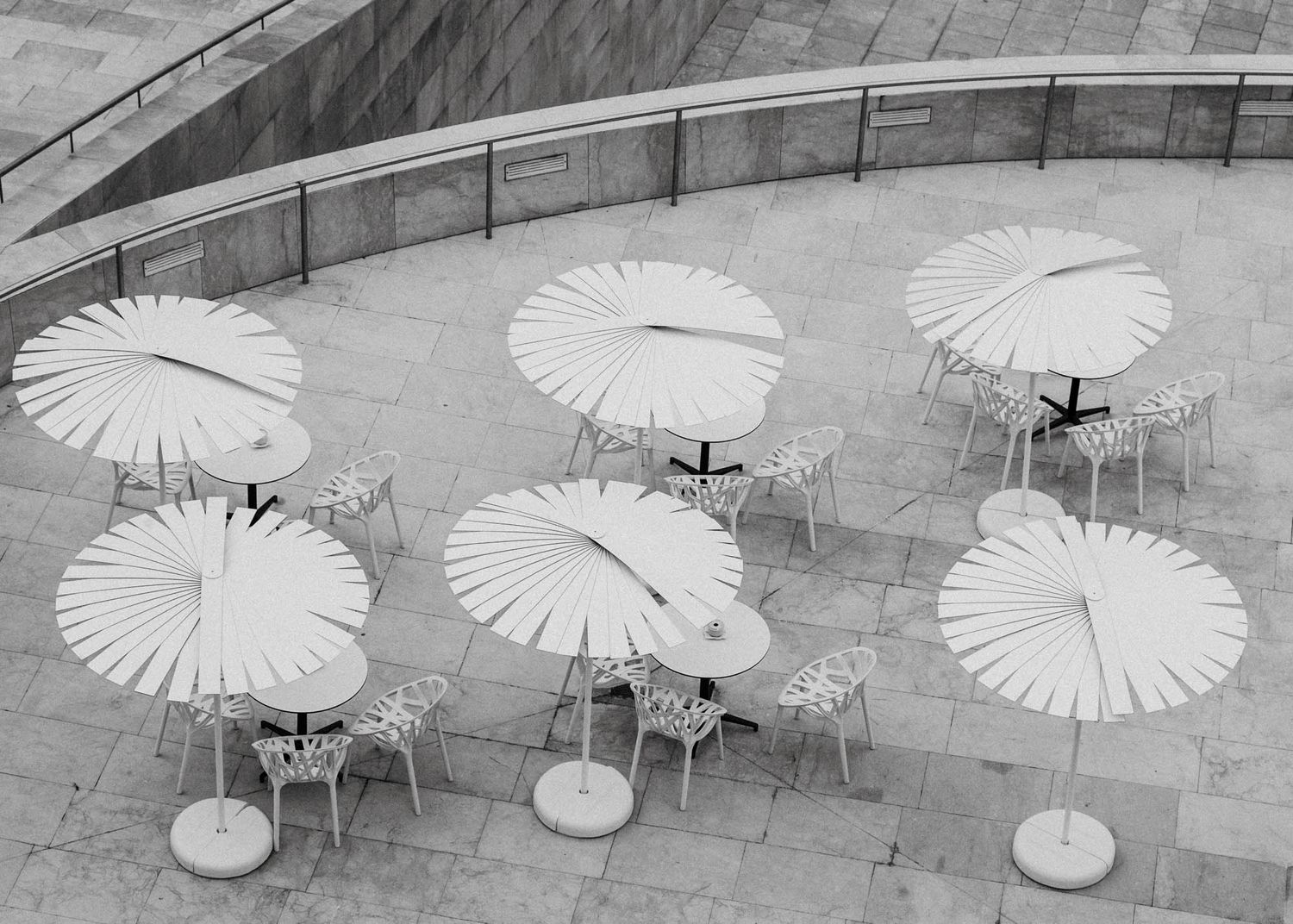 Black and white photo of white umbrellas and tables arranged on a stone patio at the Guggenheim Bilbao.