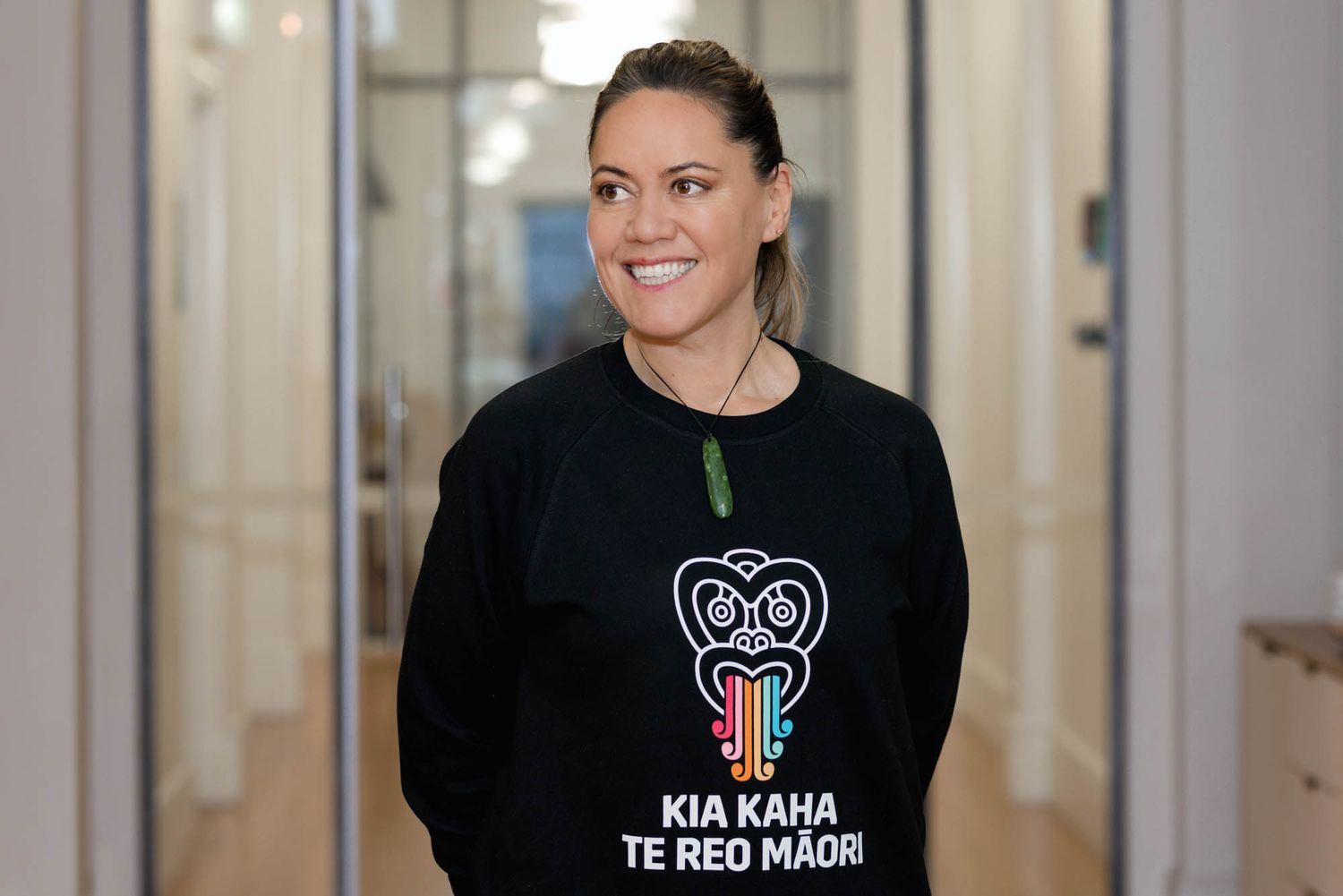 Person wearing black sweater with Maori design smiling in office hallway.