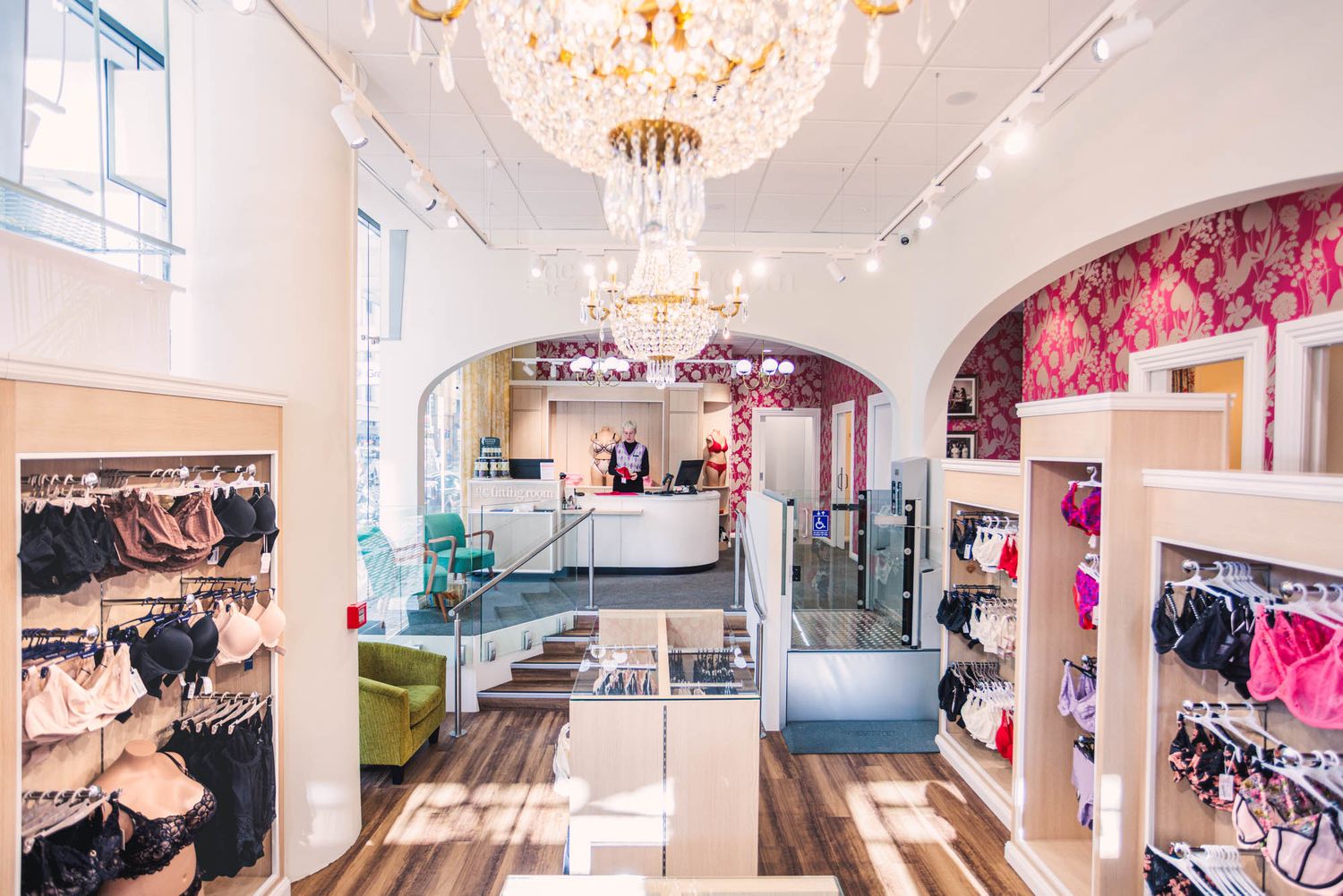 Elegant lingerie boutique interior with chandelier, arched doorways, pink wallpaper, and colorful bra displays on wooden floors.