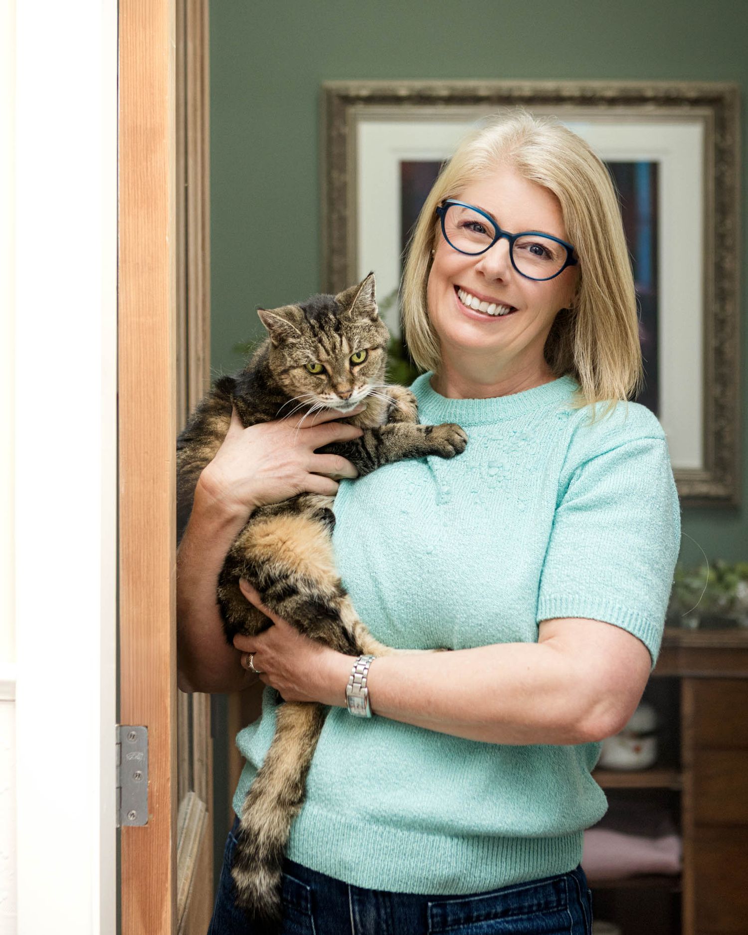 Person in mint green sweater holding a striped tabby cat while standing in a doorway.