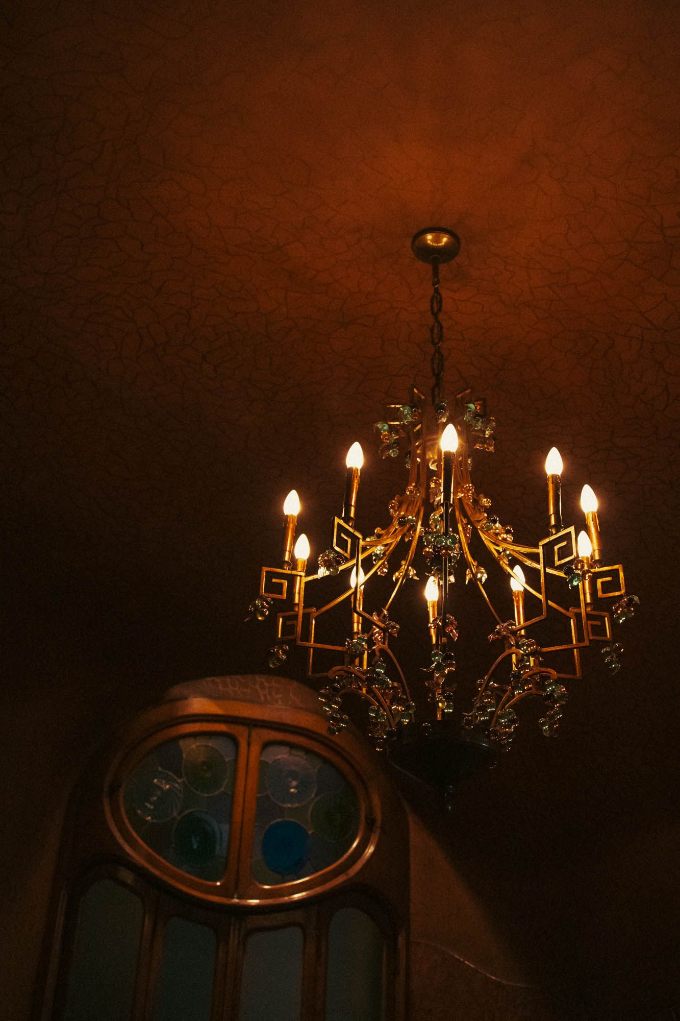 Vintage chandelier with glowing candle lights against a dark reddish-brown ceiling.