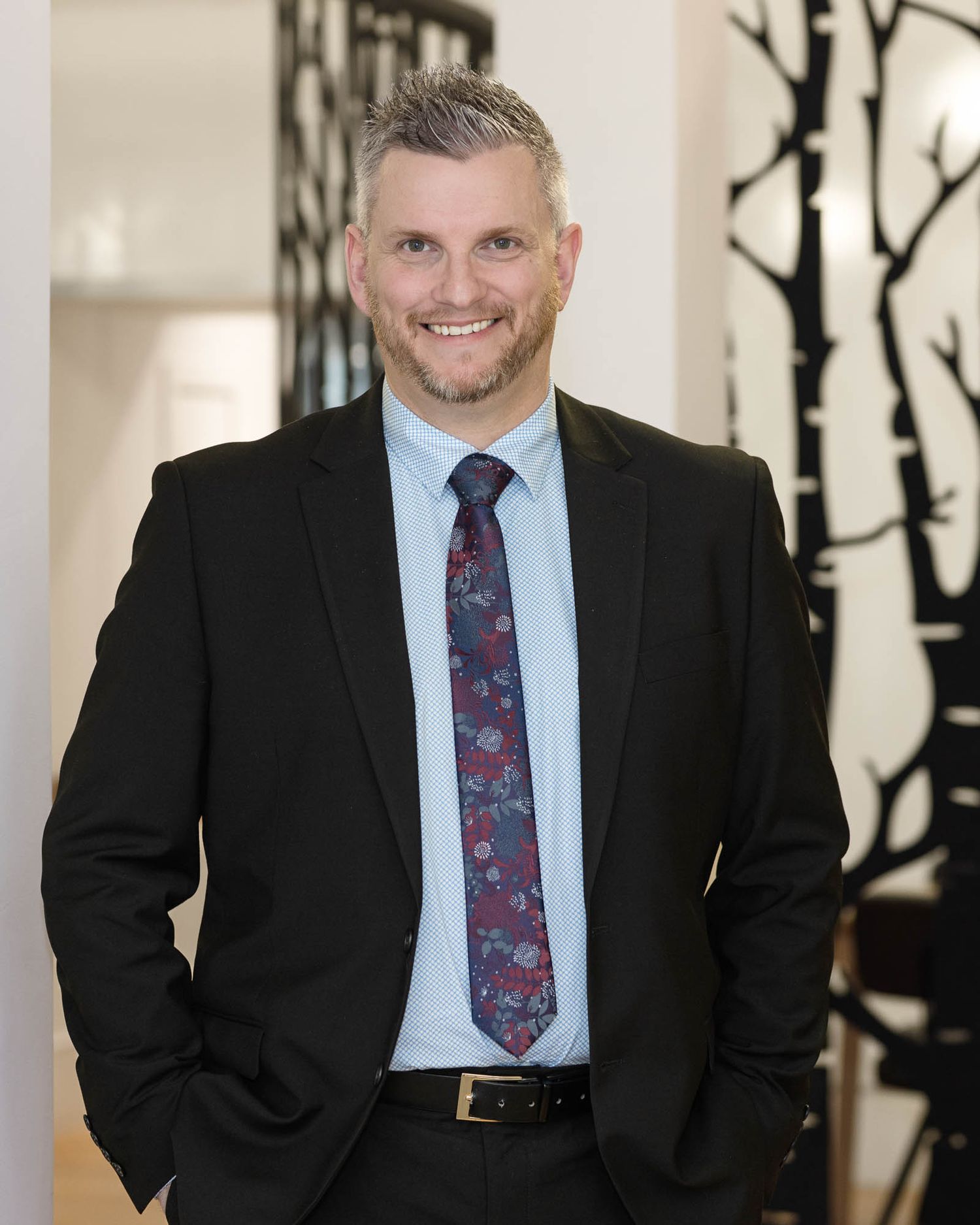 Professional man in black suit and floral tie smiling confidently in a bright modern office setting.