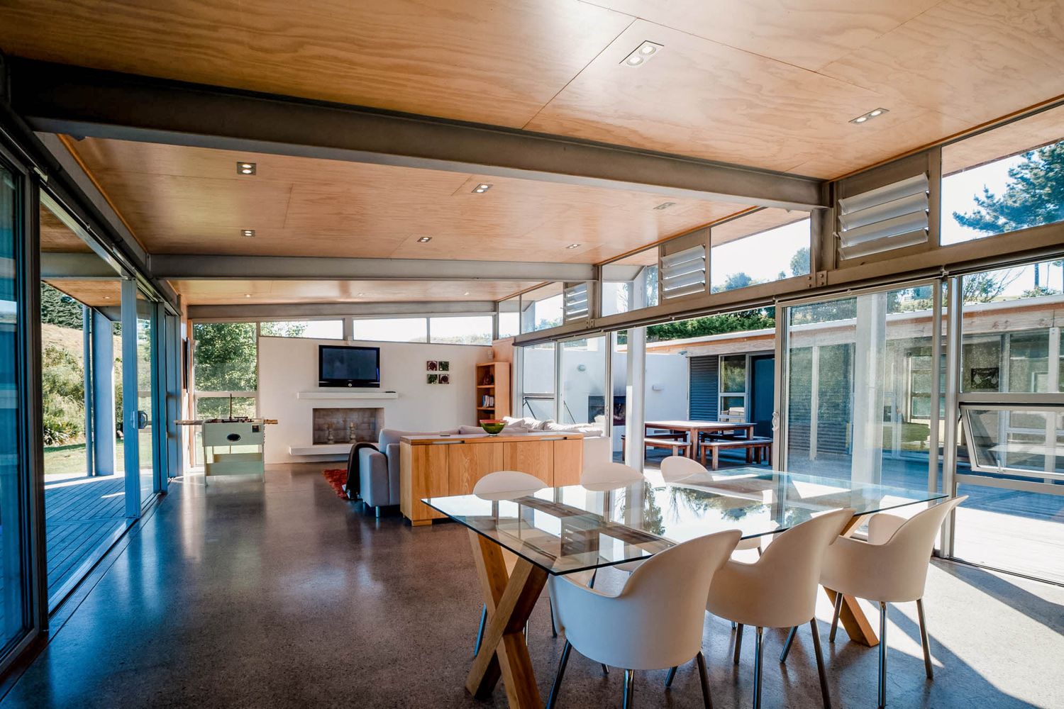 Modern open-plan dining area with glass walls, wood ceiling, white chairs, and seamless indoor-outdoor living space.