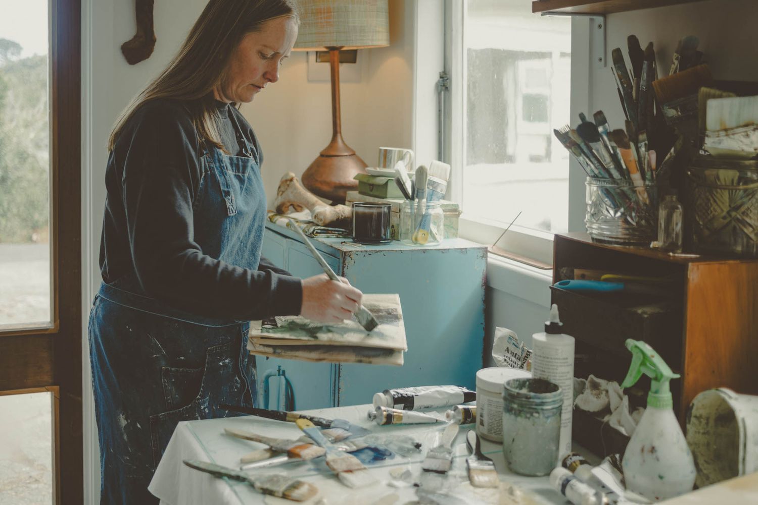 Artist working at a desk with art supplies and materials in natural light.
