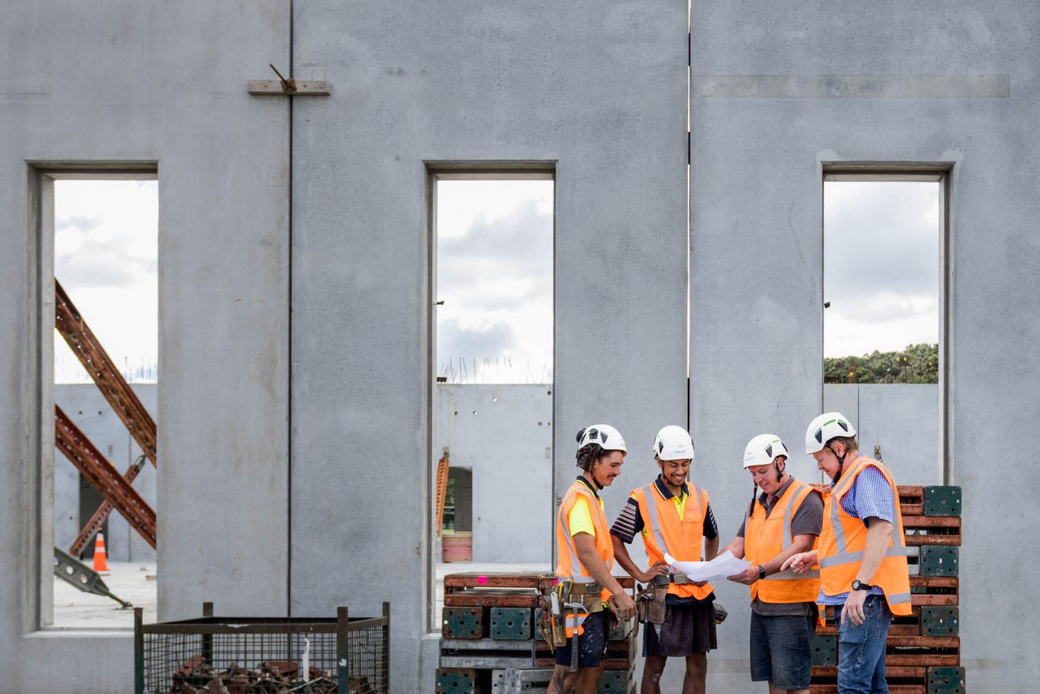 Maycroft construction workers in orange safety vests inspect a concrete wall installation at a building site.
