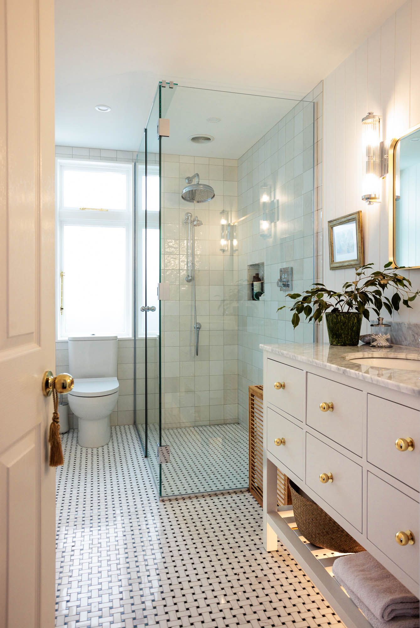Elegant white bathroom with glass shower, mosaic tile floor, white vanity with brass hardware, and a potted plant.