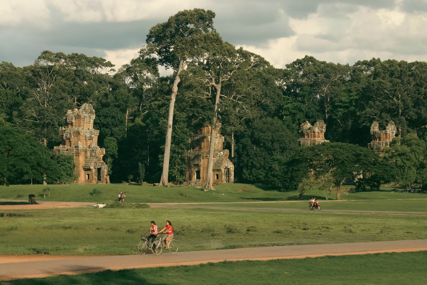 Ancient temple ruins in Angkor Wat surrounded by green lawns and trees under cloudy skies.
