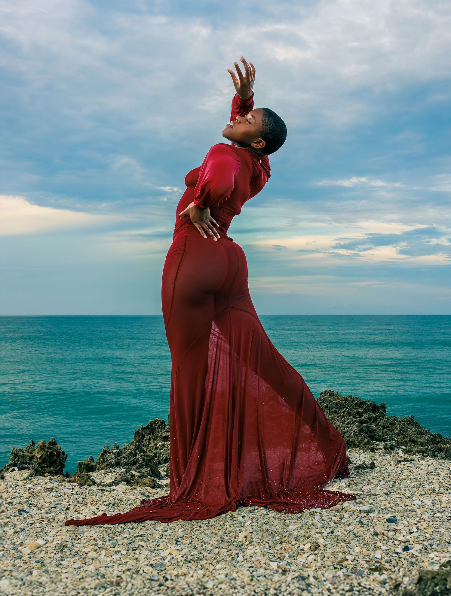 Figure in flowing red dress poses dramatically against turquoise ocean backdrop on rocky coastal terrain.