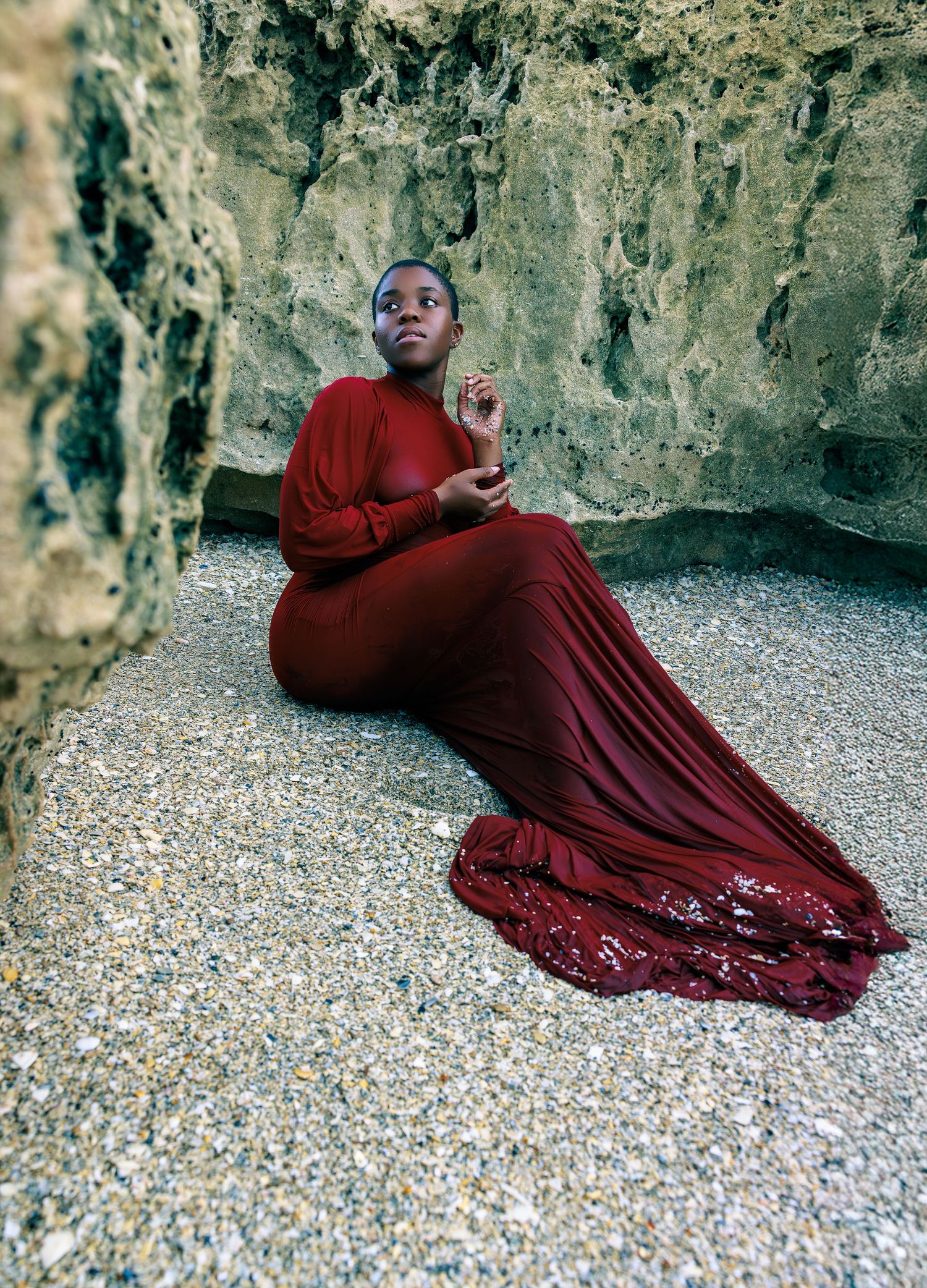 A figure in a flowing burgundy dress reclines gracefully against a rocky cave wall on a pebbly surface.