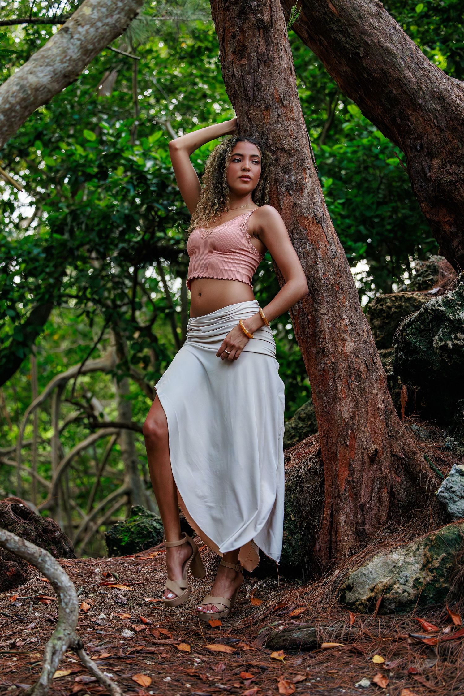 Woman in pink crop top and white slit skirt poses gracefully against a tree in a lush green forest.