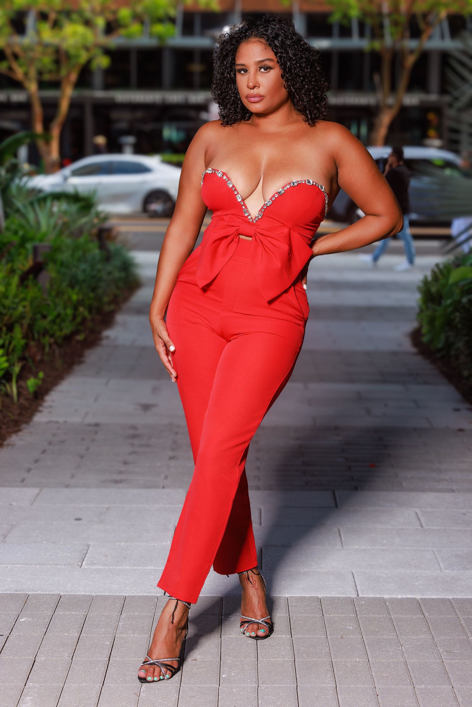 Woman in red embellished jumpsuit with sweetheart neckline poses elegantly on urban sidewalk.