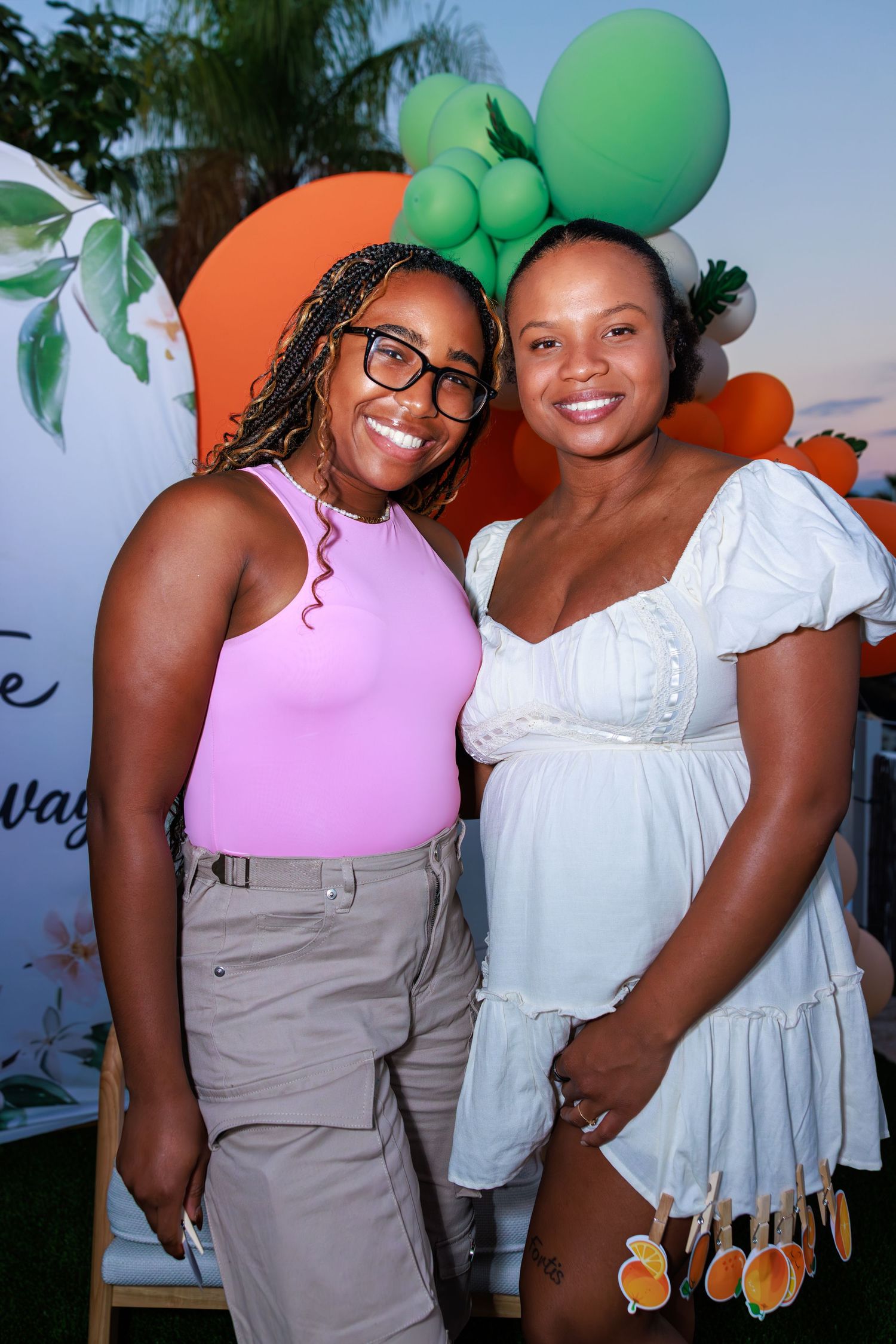 Two people wearing pink and white outfits pose together at an event with green balloons in the background.