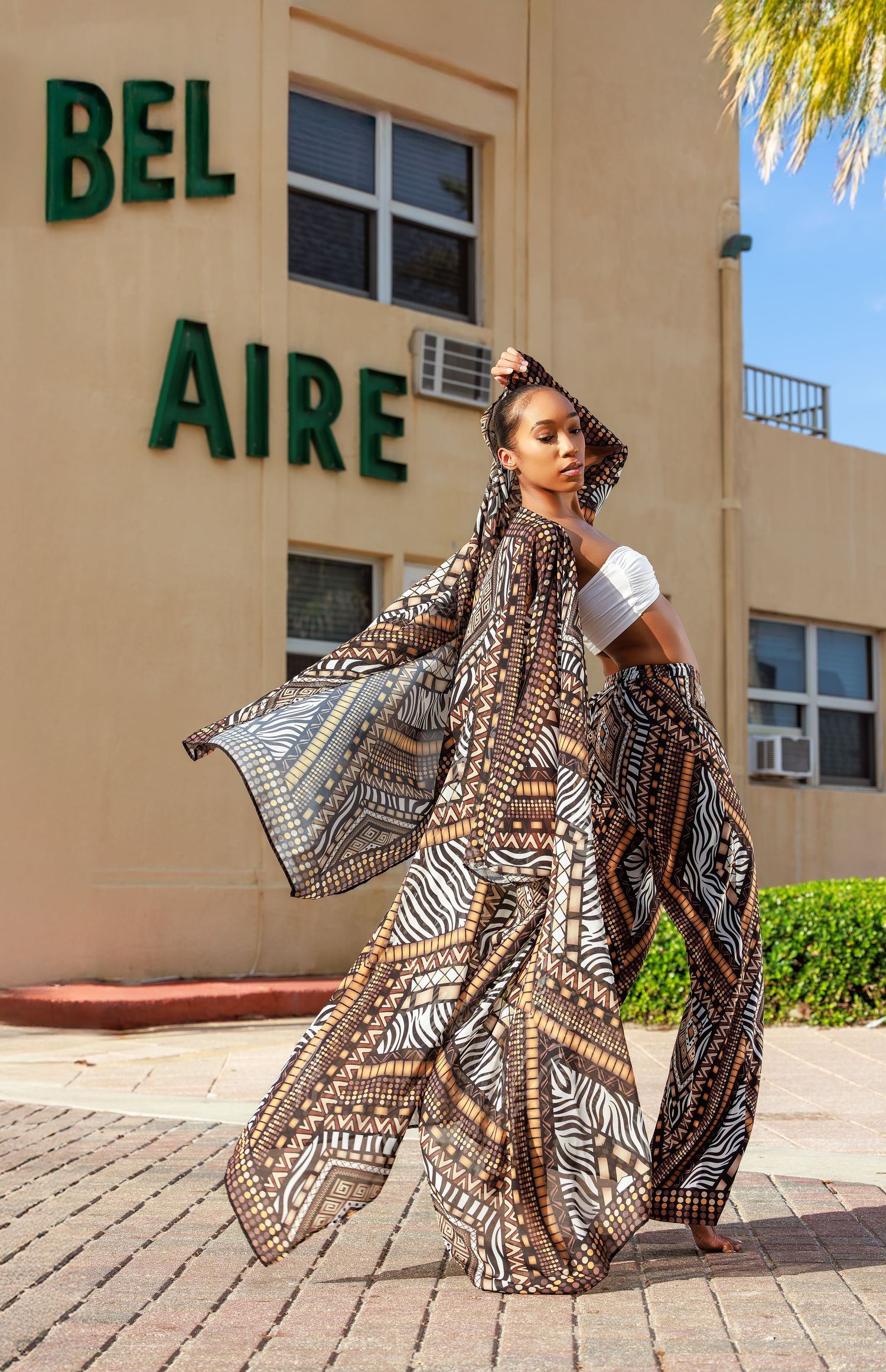 Woman posing in flowing brown tribal print kimono and wide-leg pants outside Bel Aire building on sunny day.