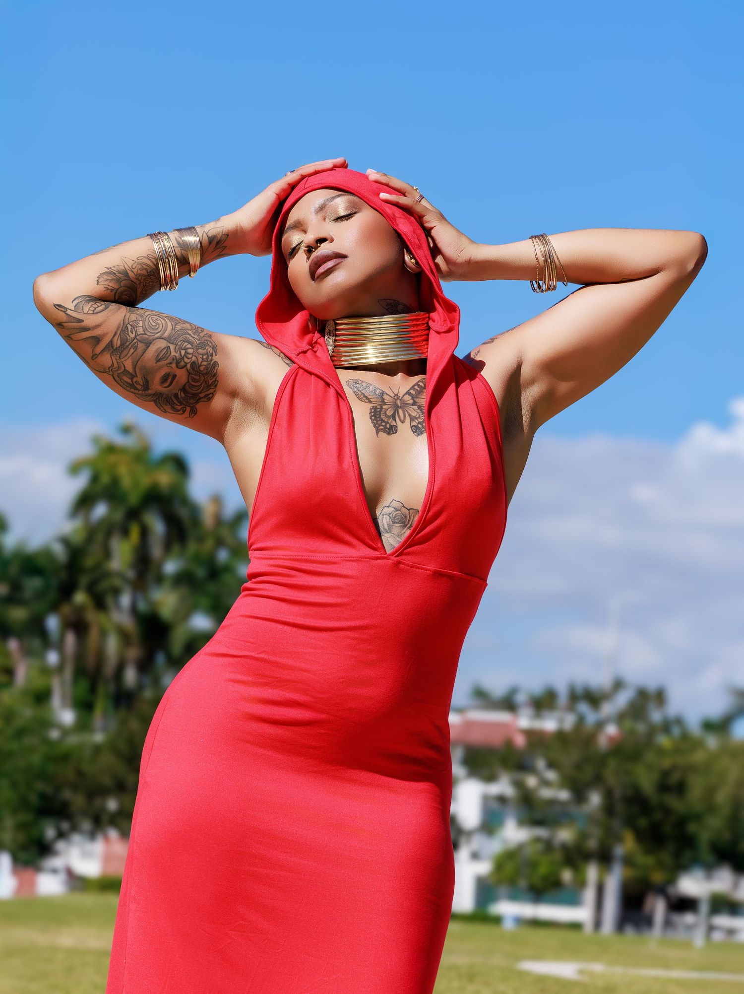 Fashion model in red plunging dress and matching headscarf poses outdoors against palm trees and blue sky.