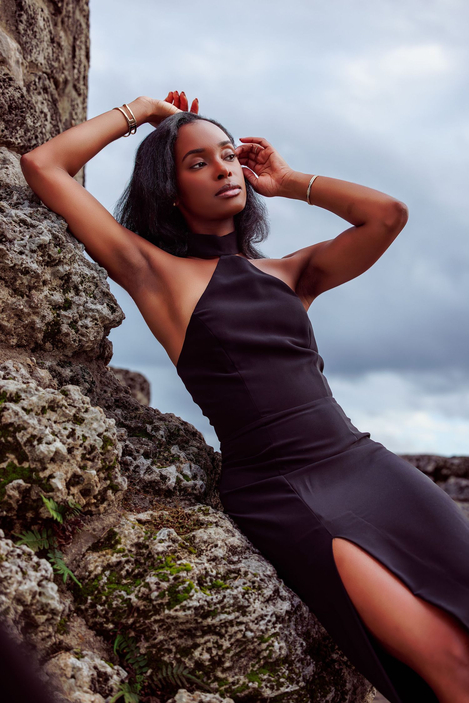 A young Black woman in a black halter dress poses gracefully against ancient stone ruins under a cloudy sky.
