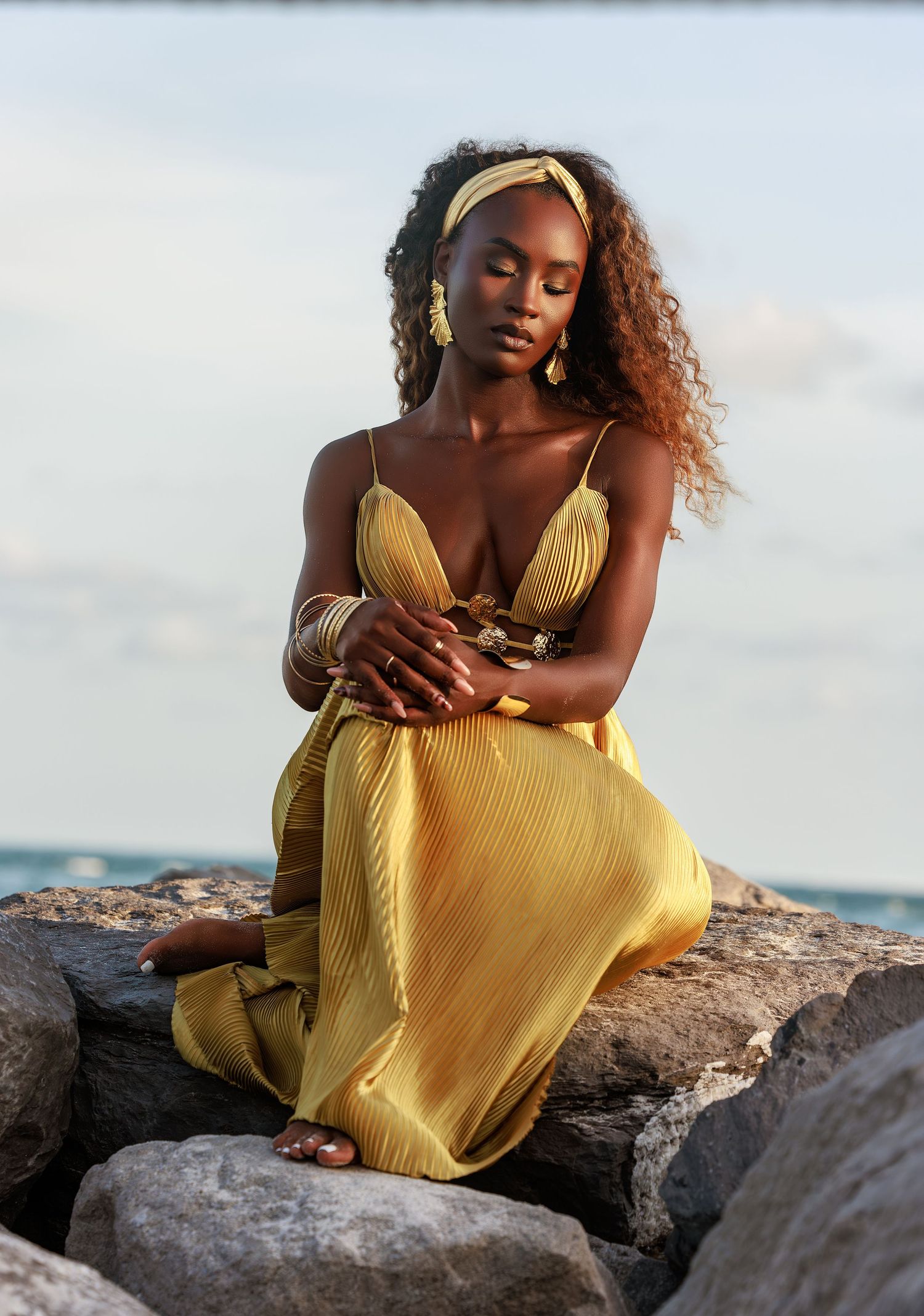 A woman in a yellow maxi dress sits on rocks by the ocean, wearing gold jewelry and a headband.