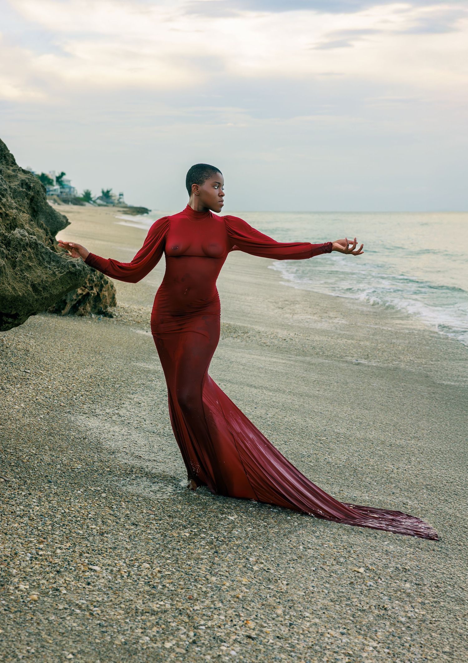 Figure in flowing red dress moves gracefully along rocky beach shoreline at sunset.