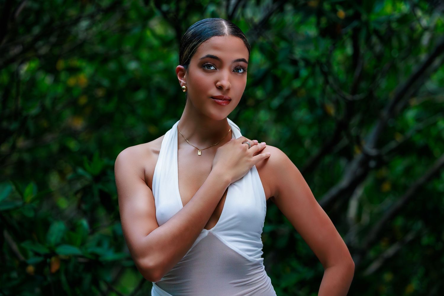 Young woman in white halter dress posing gracefully against lush green foliage backdrop.