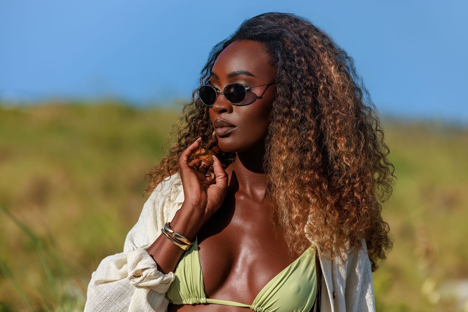 Young Black woman with curly hair wearing sunglasses and green bikini top outdoors on a sunny day.