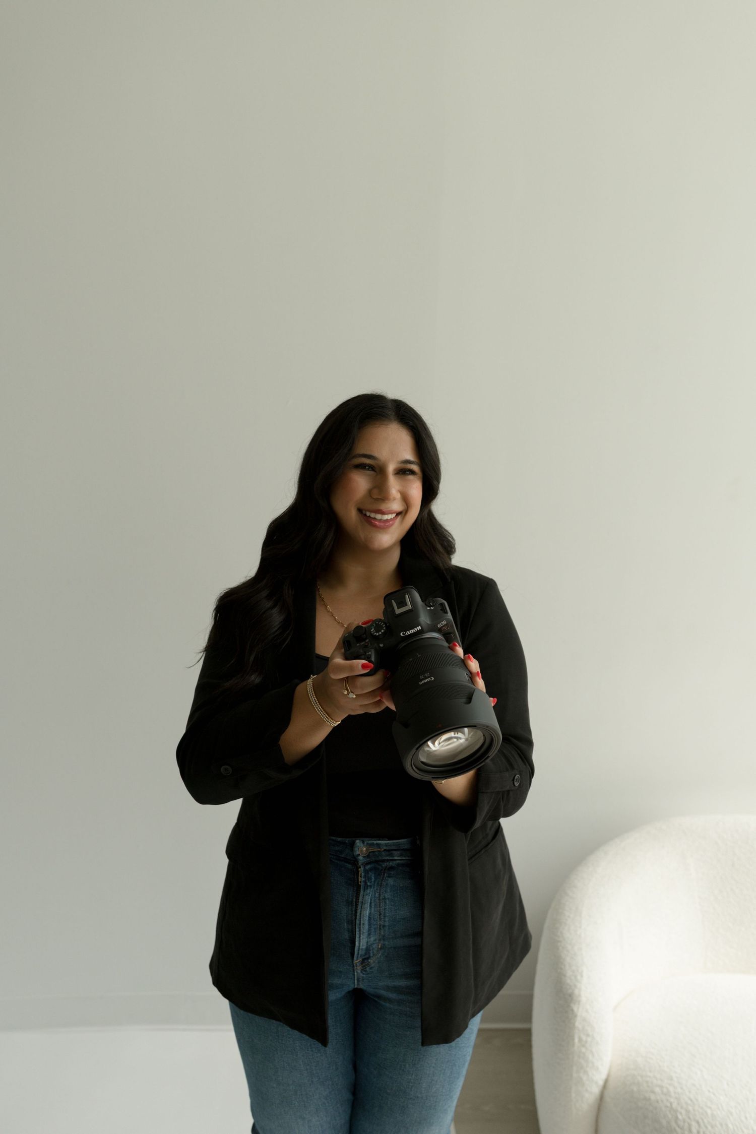 Photographer in black blazer holding camera while smiling against white background.