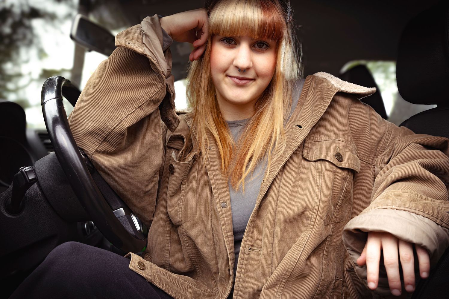 Photo of a young woman in a car, elbow on the steering wheel, turned to look at the camera. Cordouroy jacket.