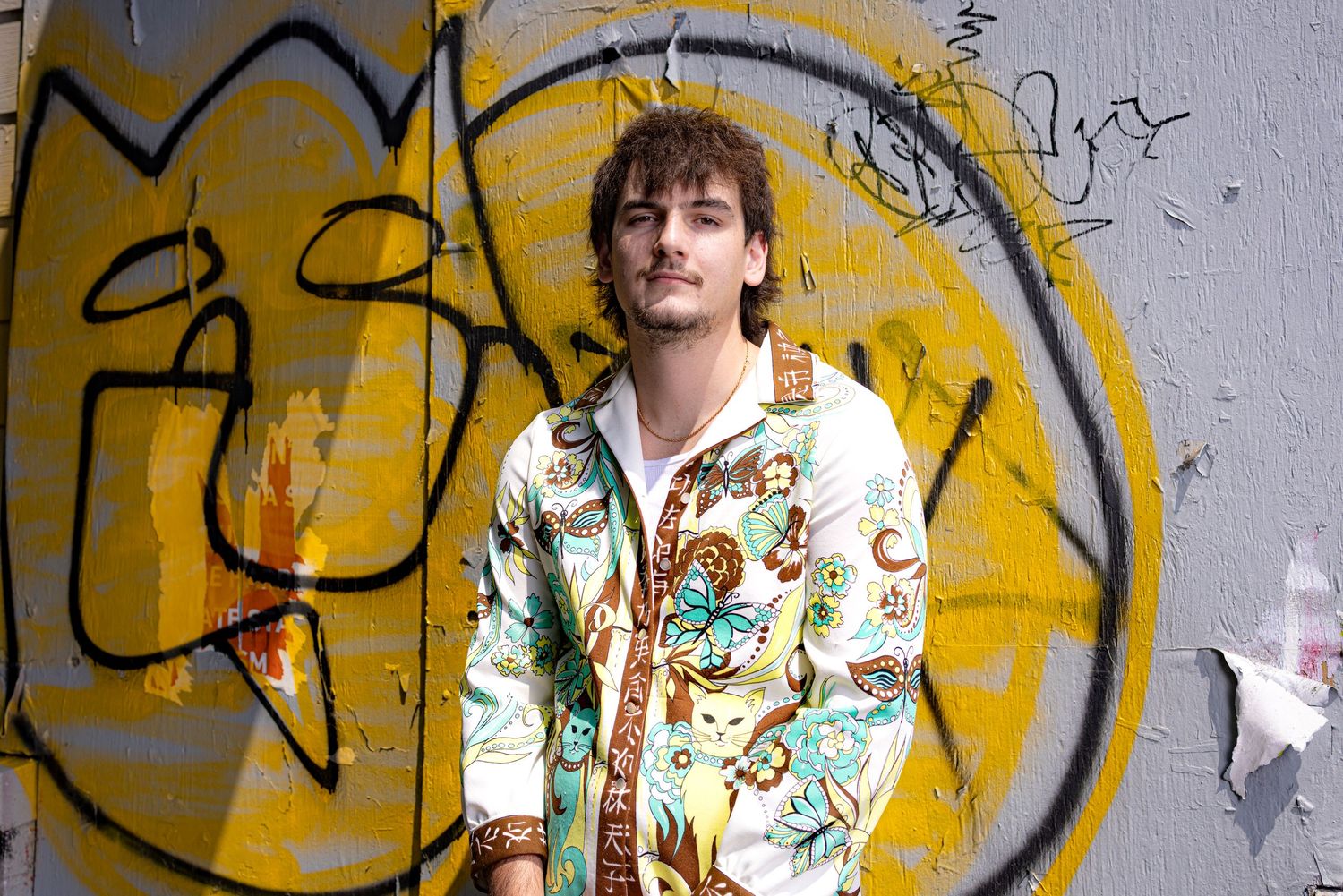 Portrait of a young man with brown hair in a dress shirt with stylized yellow & turquoise cats. Standing in front of yellow graffitied wall