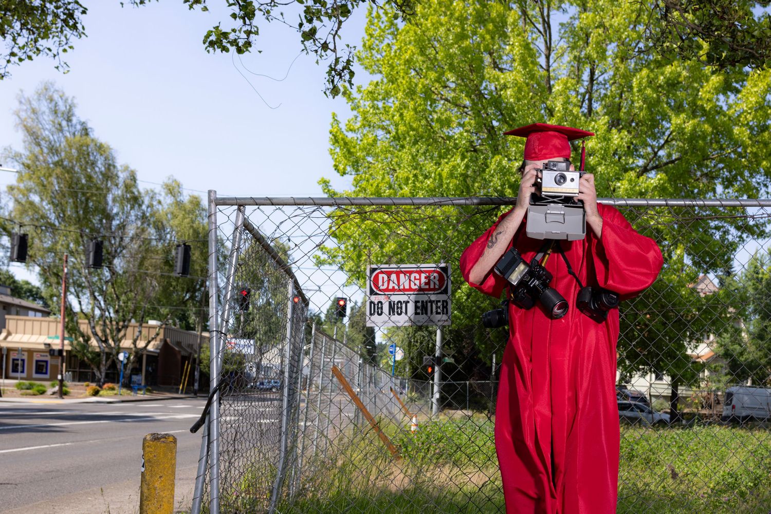 Photo of a man in a red graduation robe and cap holding a camera to his face against a chainlink fence next to a busy street