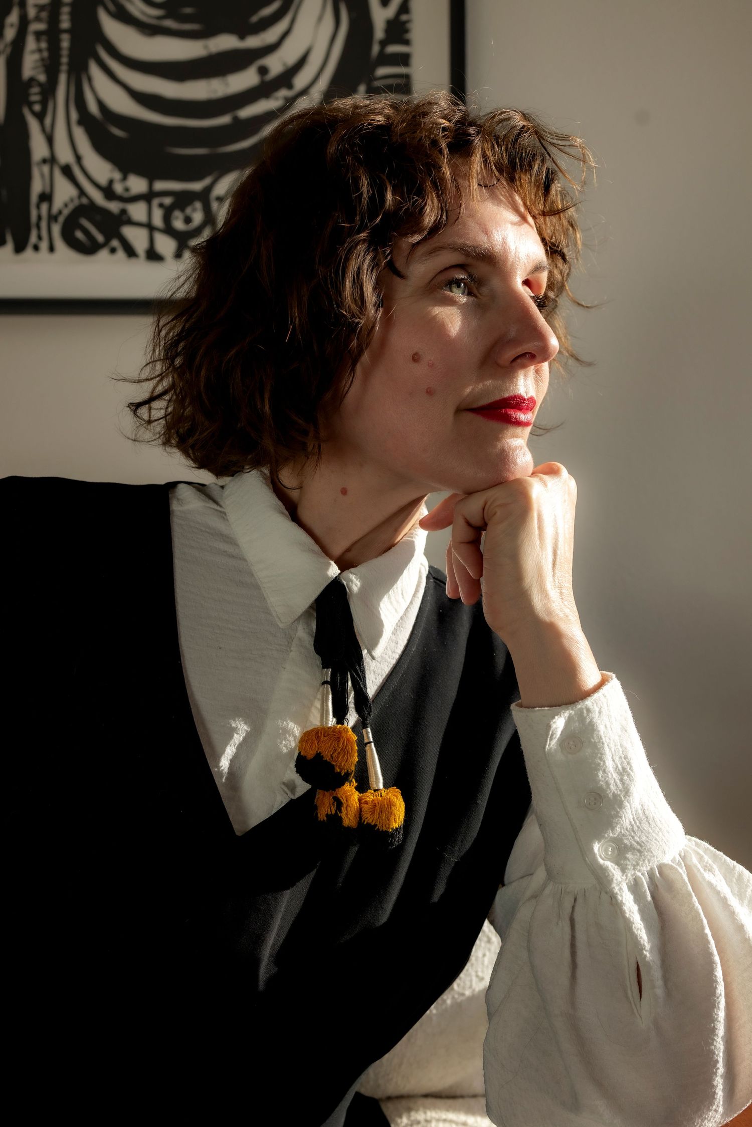 Profile portrait of a woman with brown hair, white shirt, yellow tie,and black vest looking out a window with light on one side of her face.