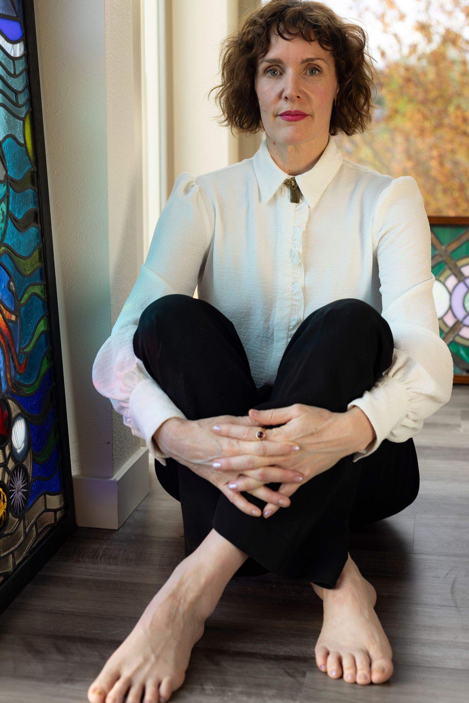 Photo of a woman with short, brown hair in a white shirt and black pants sitting cross-legged on the floor, barefoot, next to a window.