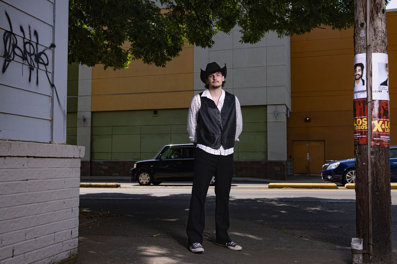 Ful-length photo of a young man with cowboy hat and vest standing on a street corner with cars driving behind him