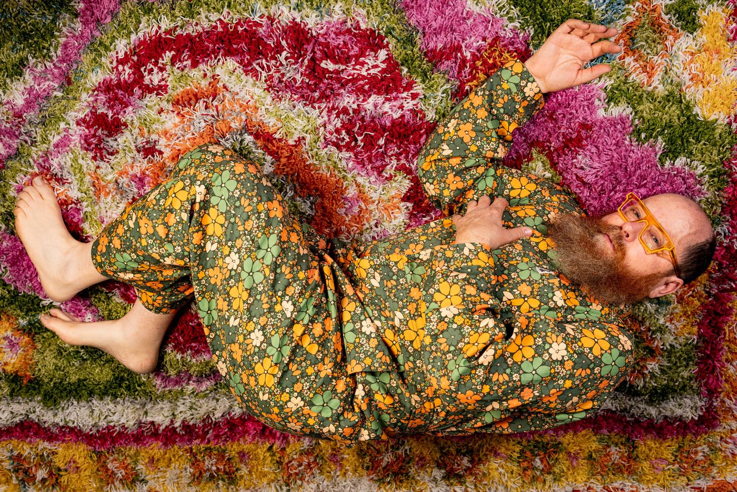 Photo of a man with orange glasses & red beard wearing a flower jumpsuit and lying on a rainbow-hued rug