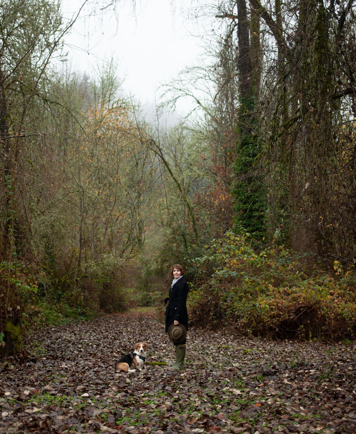 Wide photo of a woman in a trenchcoat and a corgi dog outside in the forest