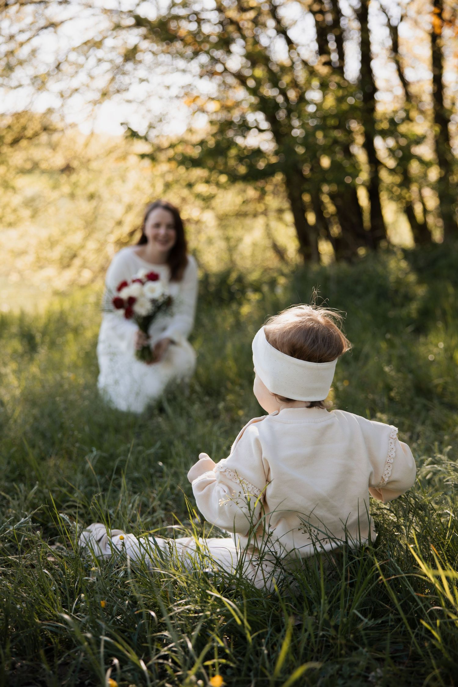 Kleinkind sitzt im hohen Gras und blickt zur Mutter, die im Hintergrund mit Blumen unscharf zu sehen ist – natürliche Familienbilder.