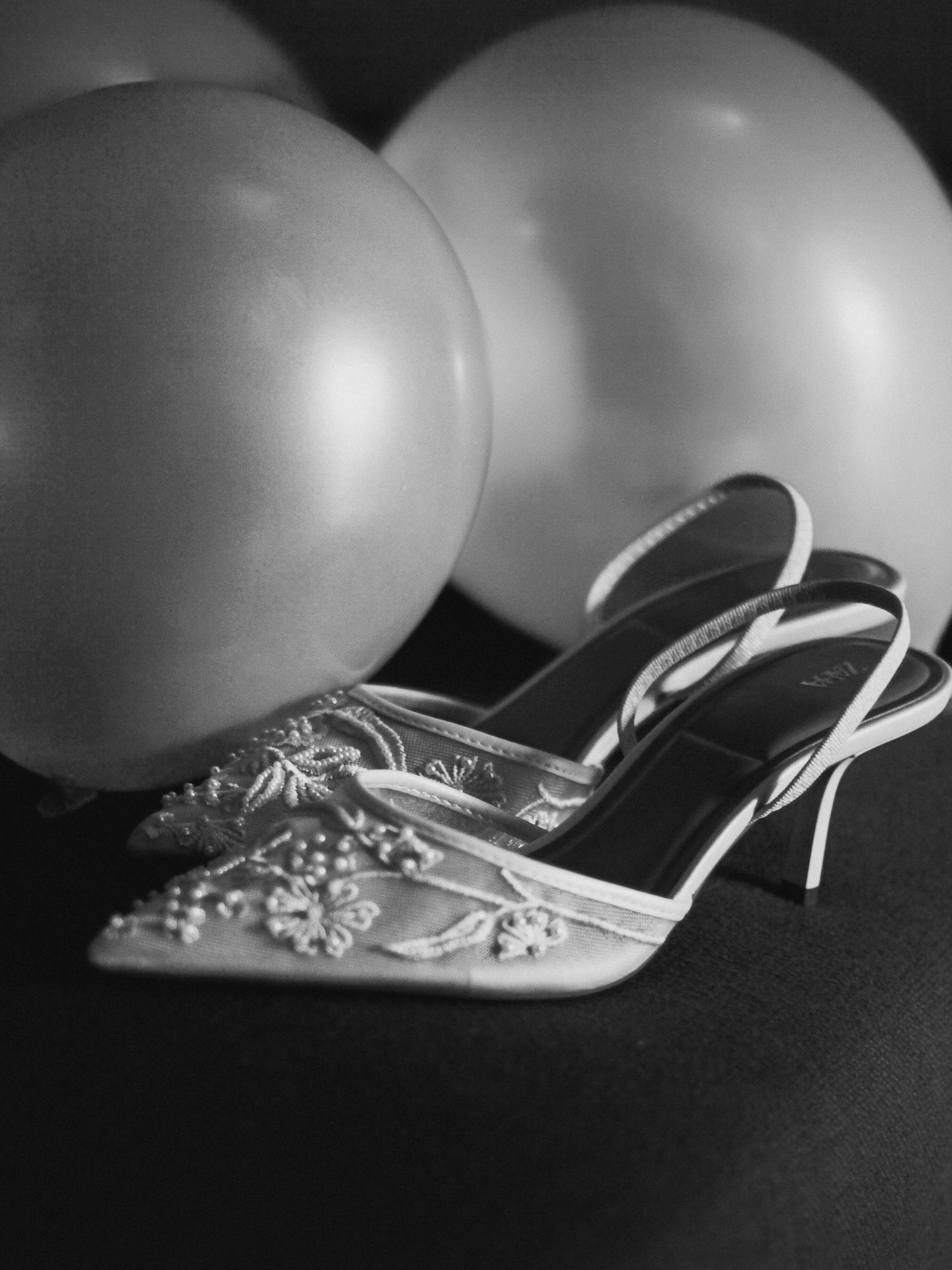 Elegante Hochzeitsdetails in Schwarz-Weiß: Bestickte Brautschuhe mit Perlenverzierung vor festlichen Luftballons im Resort.