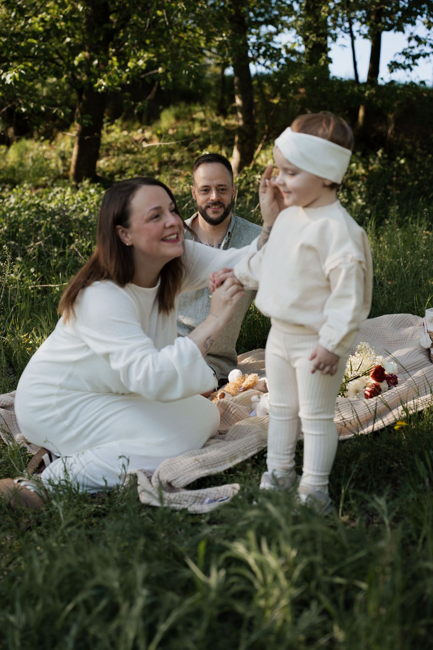 Authentisches Familienshooting in Braunau am Inn: Mutter, Vater und Kleinkind lachen gemeinsam auf einer Picknickdecke im hohen Gras bei Son