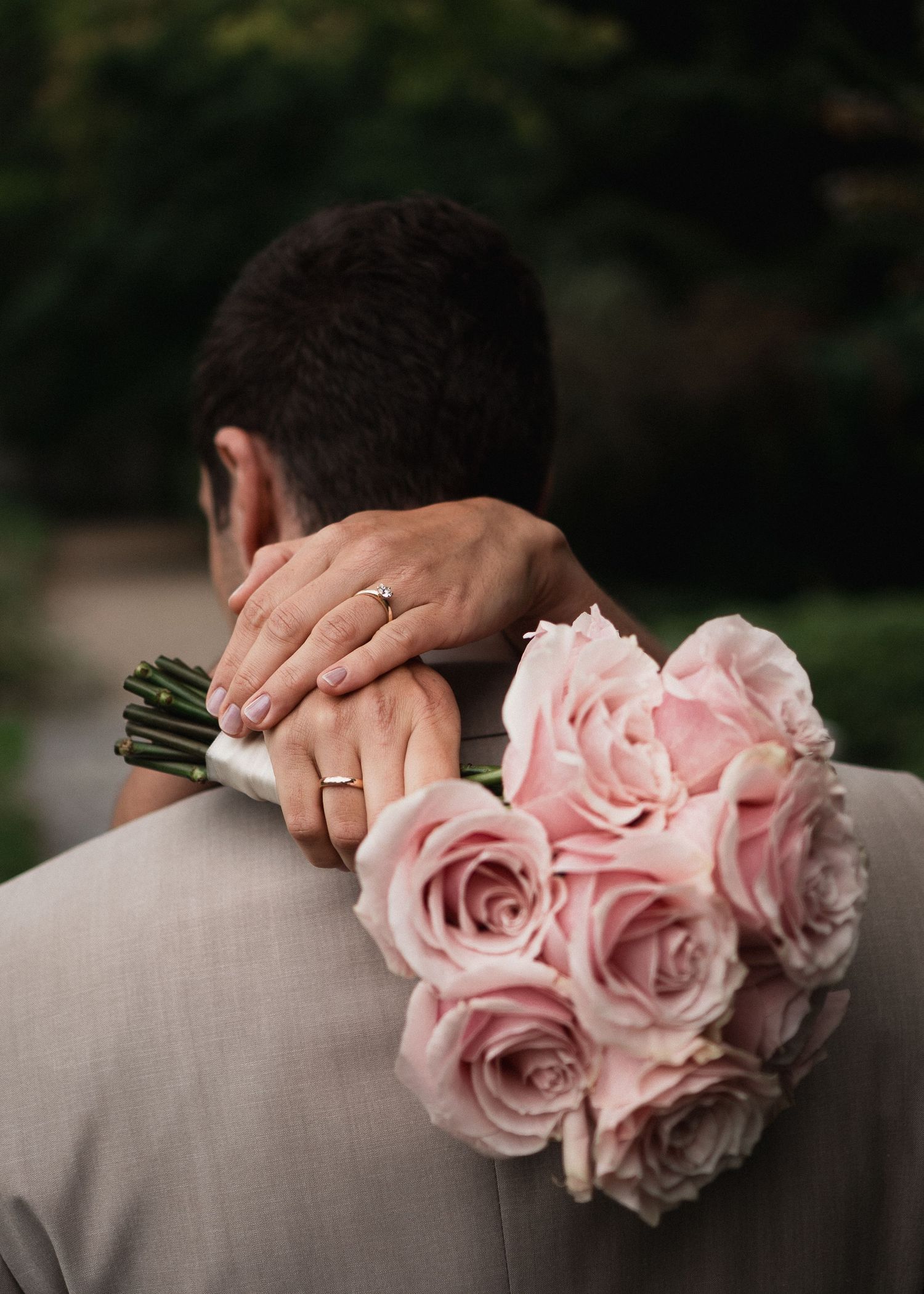Detailaufnahme der Hochzeitsringe: Die Hände der Braut ruhen auf der Schulter des Bräutigams, im Vordergrund ein eleganter Strauß aus rosa R
