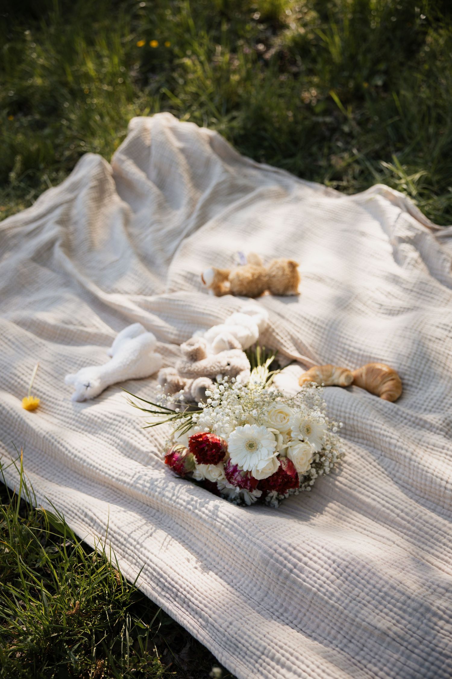 Detailaufnahme einer Picknick-Szene mit Blumenstrauß, Croissants und Kuscheltieren auf einer Decke im Gras.