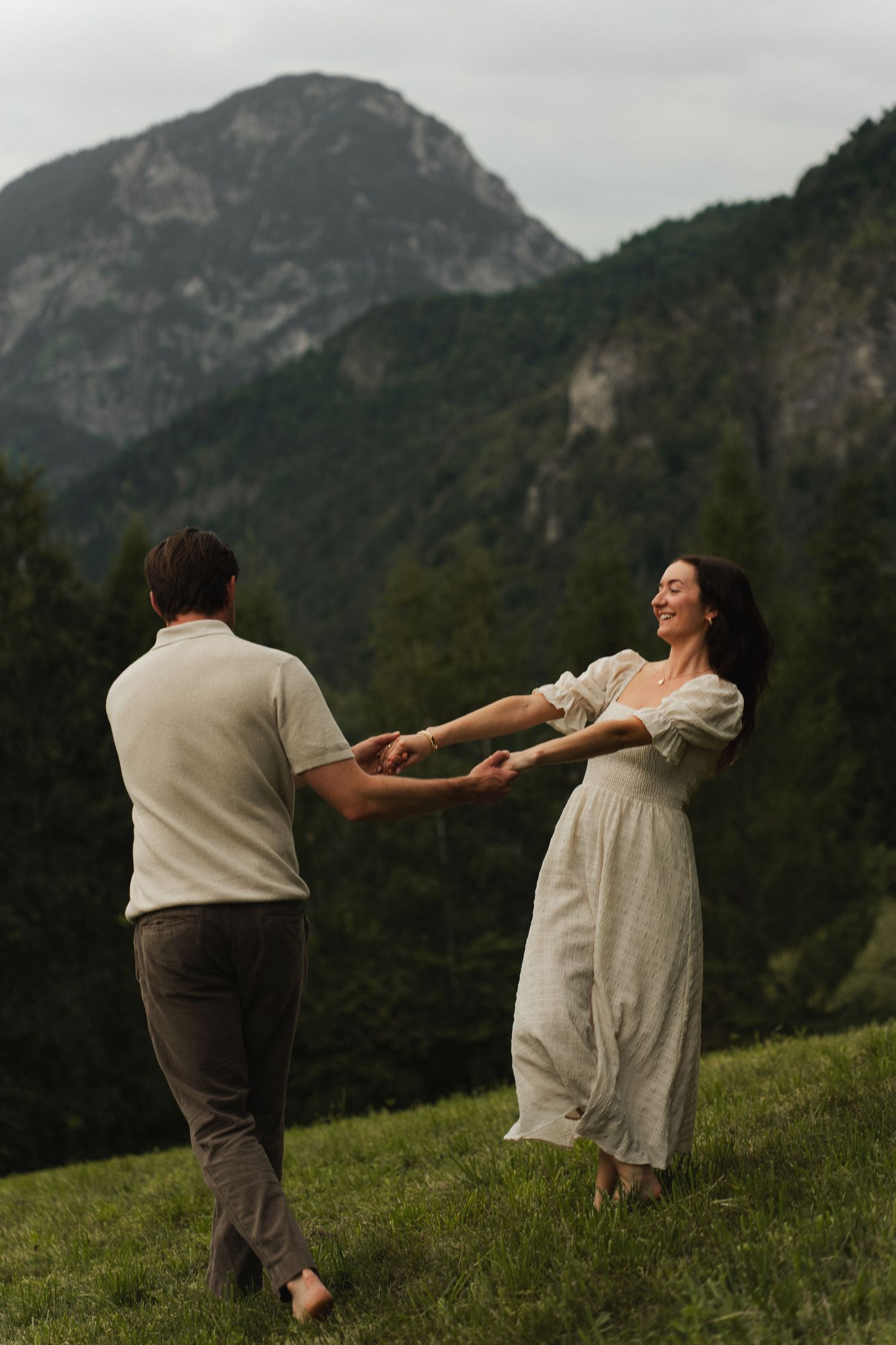 Eine cinematische Aufnahme eines Paares, das sich an den Händen hält und auf einem Feld am Thumsee vor alpiner Kulisse tanzt.
