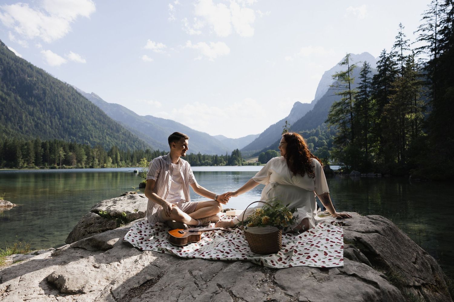 Ein junges Paar sitzt bei einem Picknick auf einem Felsen am Hintersee in den Bergen, hellen Fine-Art-Stil Ella Grinvalde Photography