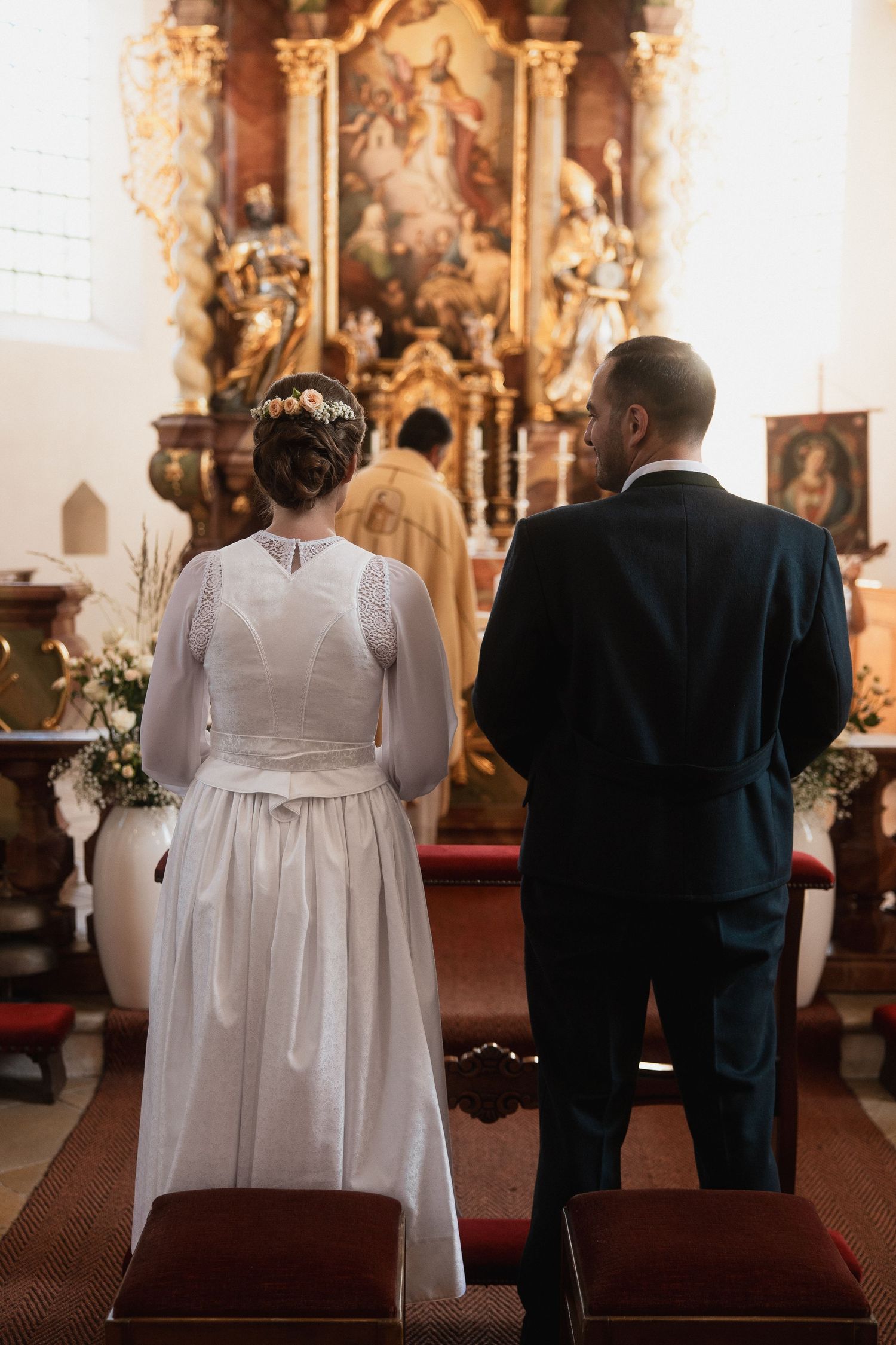 Kirchliche Trachten-Hochzeit in Bad Griesbach: Brautpaar in Hochzeitsdirndl und Trachtenanzug von hinten am Altar während der Trauung.