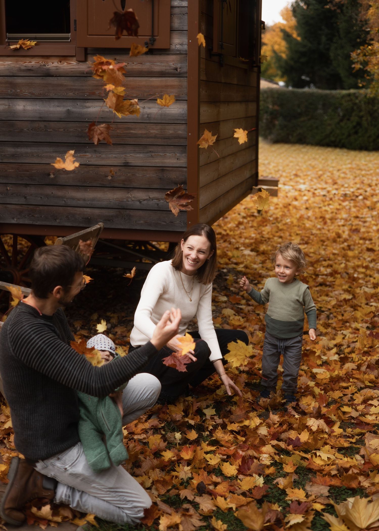 Fröhliches Herbst-Shooting am Arterhof: Eine Familie spielt mit fallenden bunten Blättern vor einem gemütlichen Holzwagen.