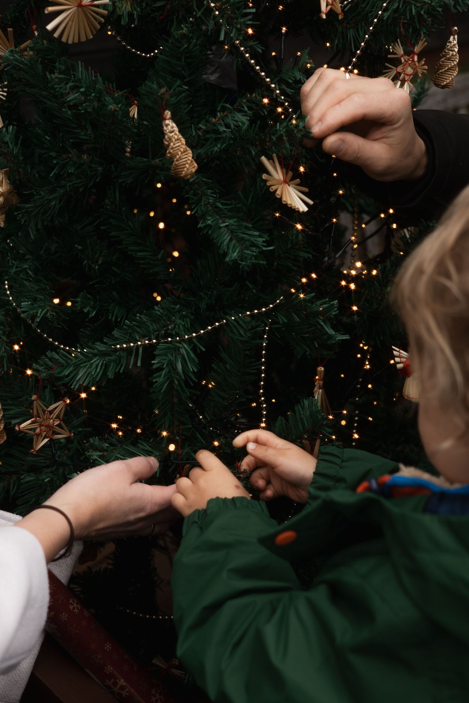 Detailaufnahme eines Kindes, das zusammen mit Eltern einen Weihnachtsbaum mit Strohsternen und Lichterketten im Arterhof schmückt.