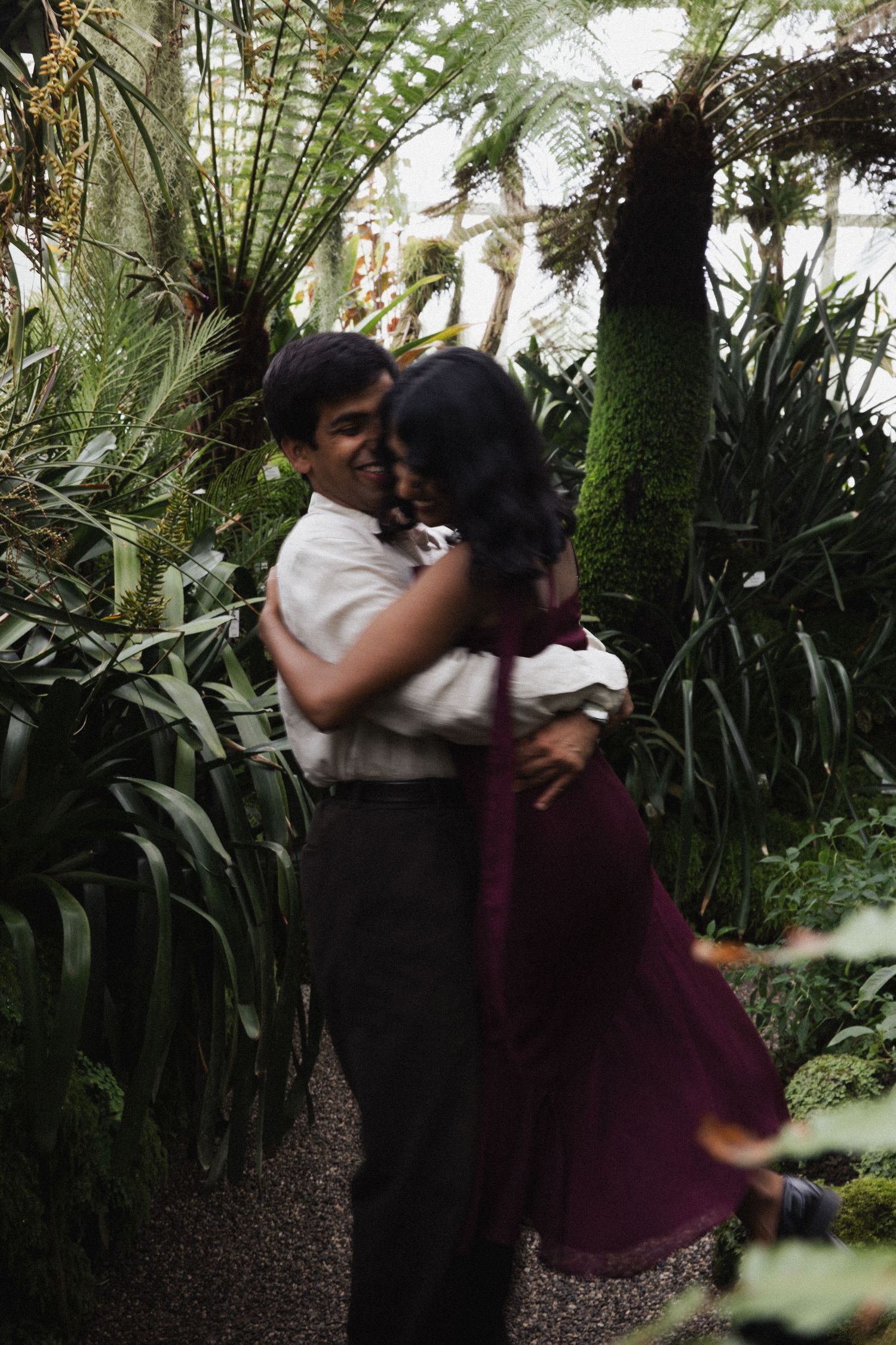 Couples Shoot in München: Paar umarmt sich voller Freude im Farnhaus des Botanischen Gartens. Dynamische, emotionale Verlobungsfotos.